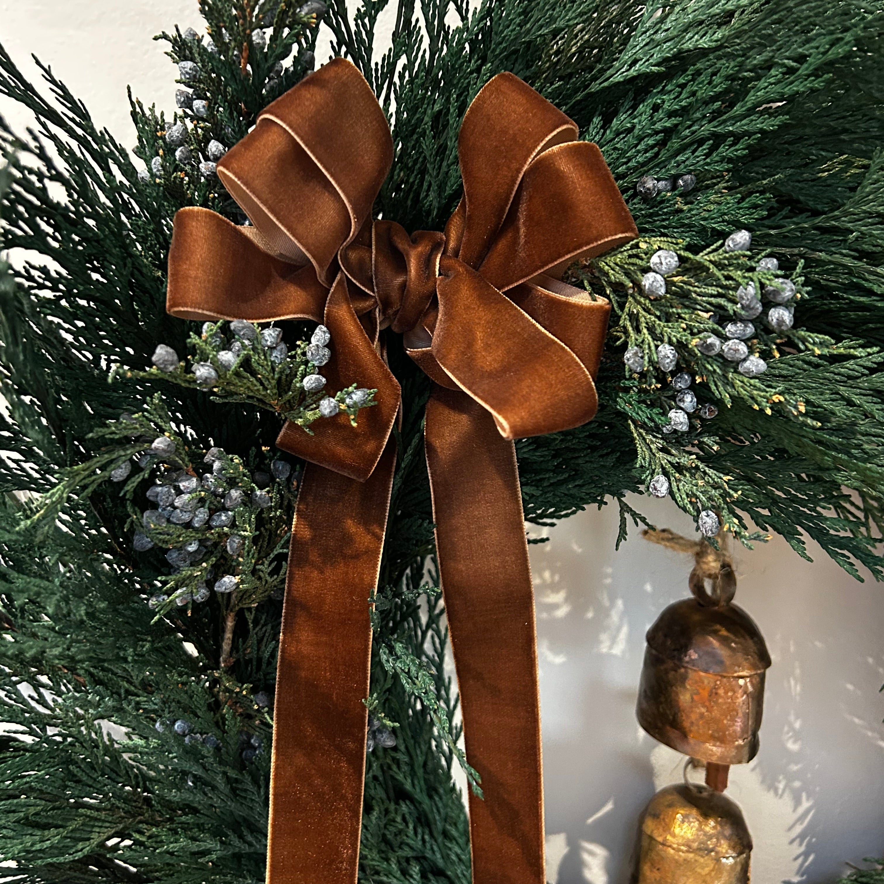 A close up ofa chocolate brown velvet bow with ribbon ends dangling, on a wreath layered with lush, preserved Cedar and accented with vibrant Juniper Berries,  and a pair of iron bells on jute twine hanging in the middle.