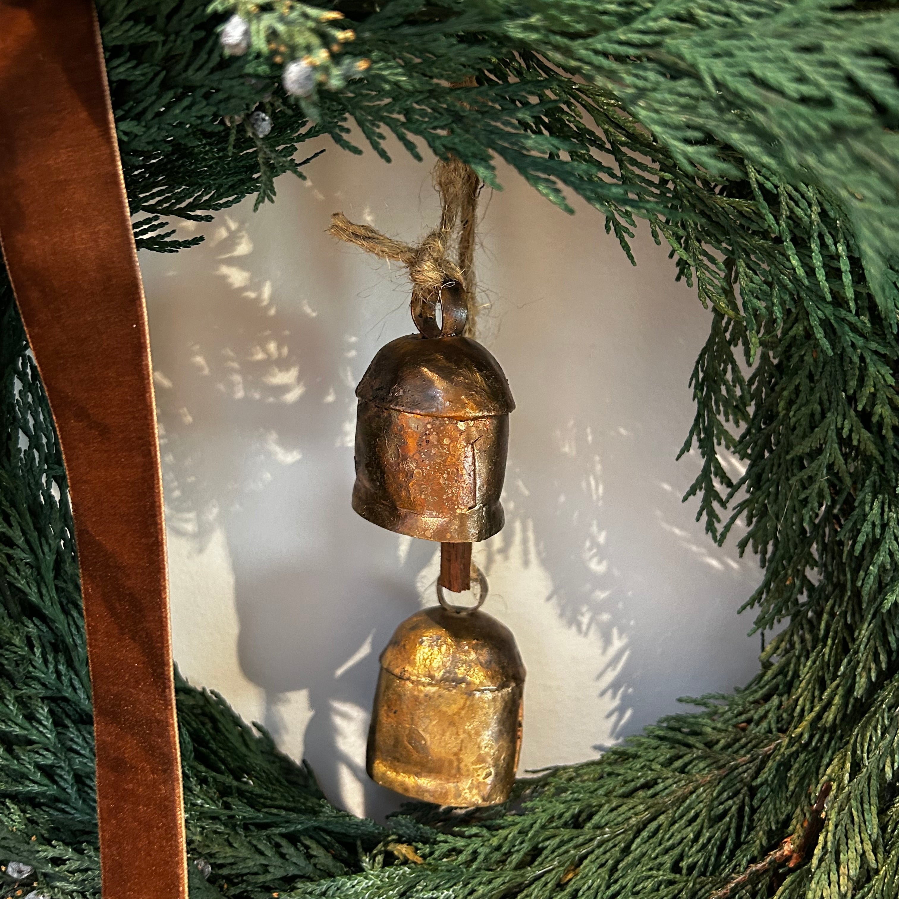 Close up of a wreath layered with lush, preserved Cedar and accented with vibrant Juniper Berries, a chocolate brown velvet bow with ribbon ends dangling, and a pair of iron bells on jute twine hanging in the middle.