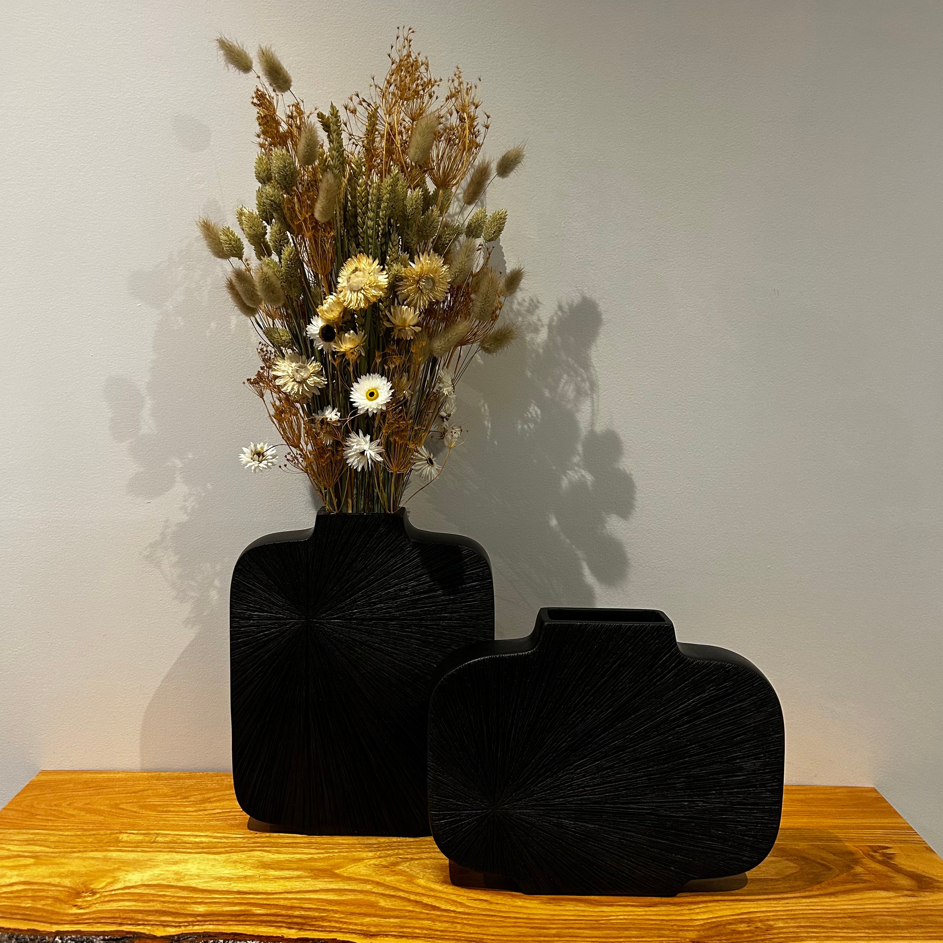 Two striking, sculptural vases in midnight-cocoa with a bold, radiating carved pattern that starting in the left corner. The tall version of the vase is hosting on of our beautiful bouquets, while the wide version of the vase is empty.