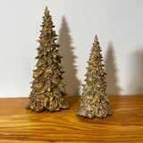 Two gold Christmas tree figurines on a wooden surface with a white background