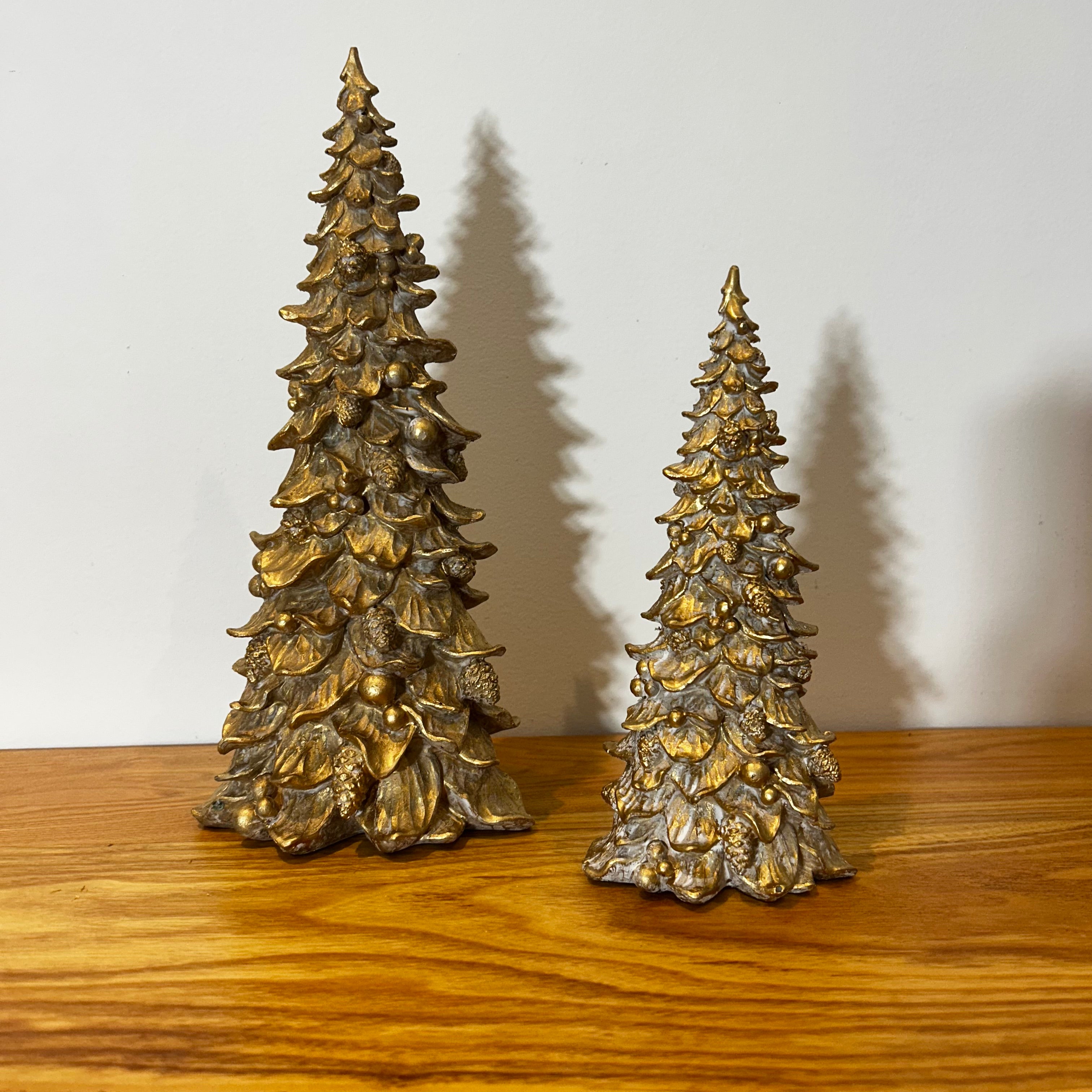 Two gold Christmas tree figurines on a wooden surface with a white background