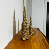 Side view of two gold Christmas tree figurines on a wooden surface with a white wall background.