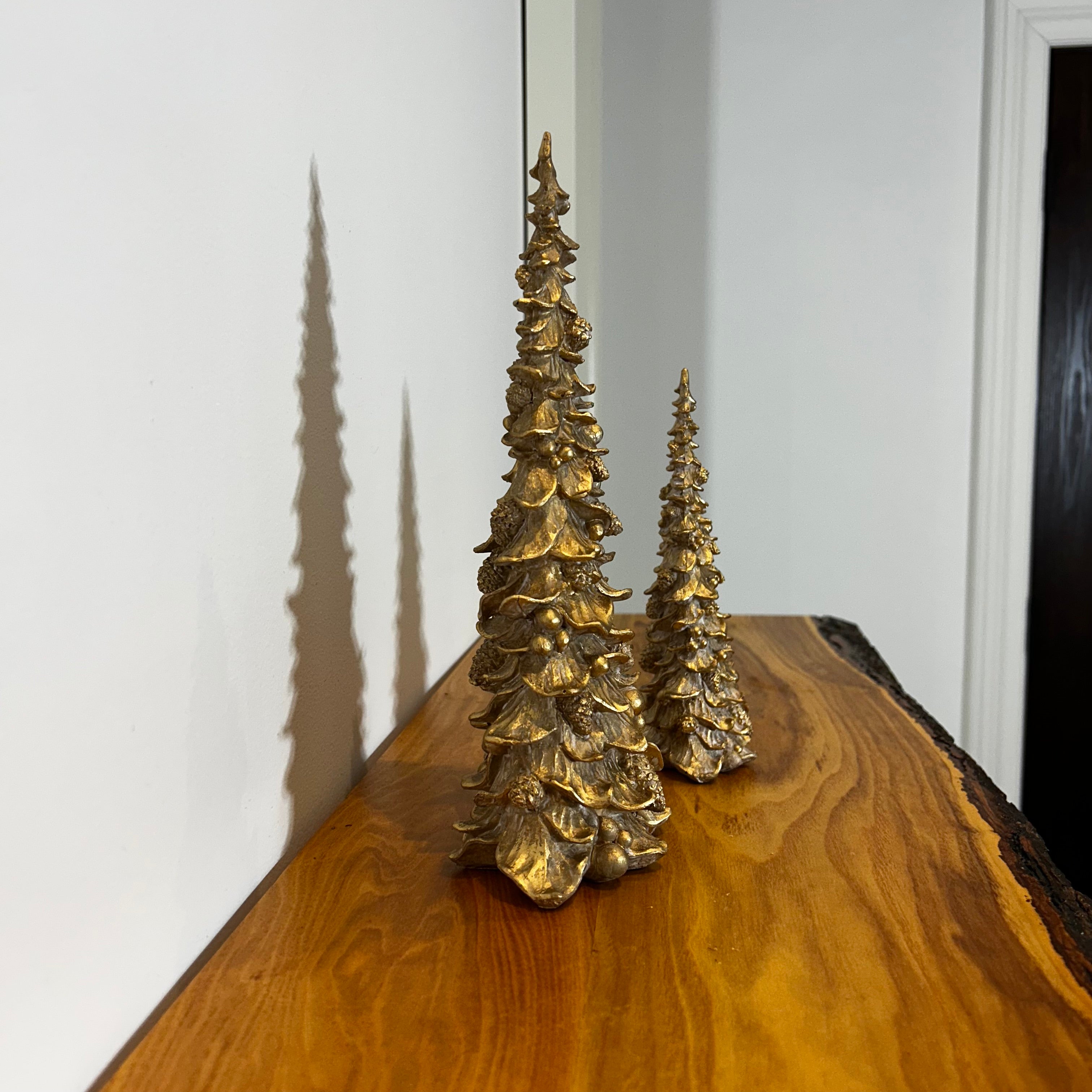 Side view of two gold Christmas tree figurines on a wooden surface with a white wall background.