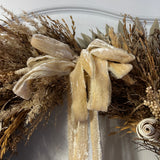 A close-up of the ragged edged velvet bow on an impressive 28 inch wreath made with the elegance of spider wood and the airy lightness of Star Grass. It is a rich tapestry of grains, including the unique forms of Sorghum, dried Rice Ears, and graceful Arena Wild Oats. Sculptural bleached cane cones and the whimsical twists of bleached curly ting ting add the final, enchanting touches.