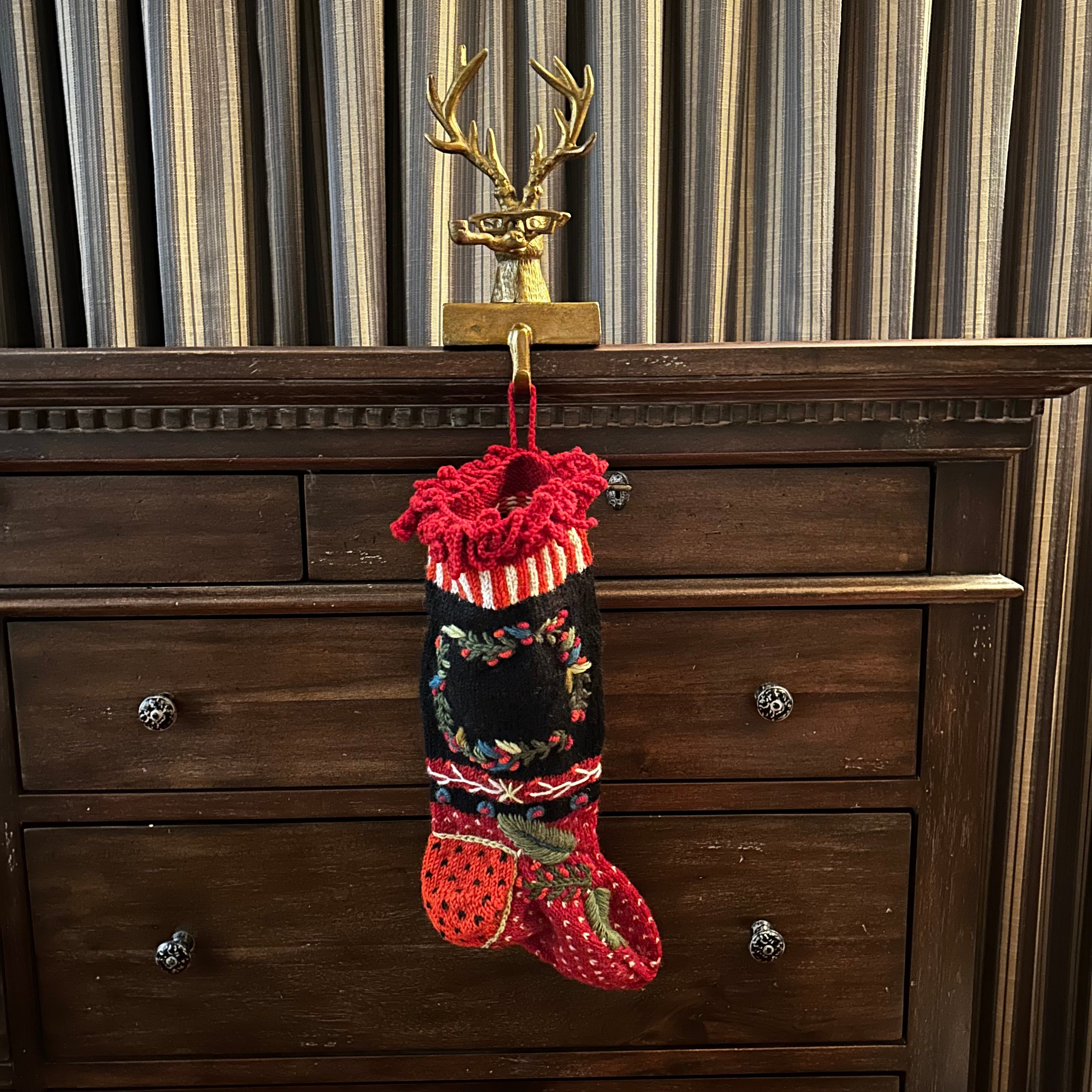 Decorative bronze deer head stocking holder hook on a wooden dresser holding a beautiful  decorative hand-knit stocking with a hand-embroidered wreath. The deer is quite a character wearing glasses and smoking a pipe. 