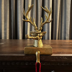 Decorative bronze deer head stocking holder hook on a wooden dresser with striped curtains in the background. The deer is quite a character wearing glasses and smoking a pipe. 