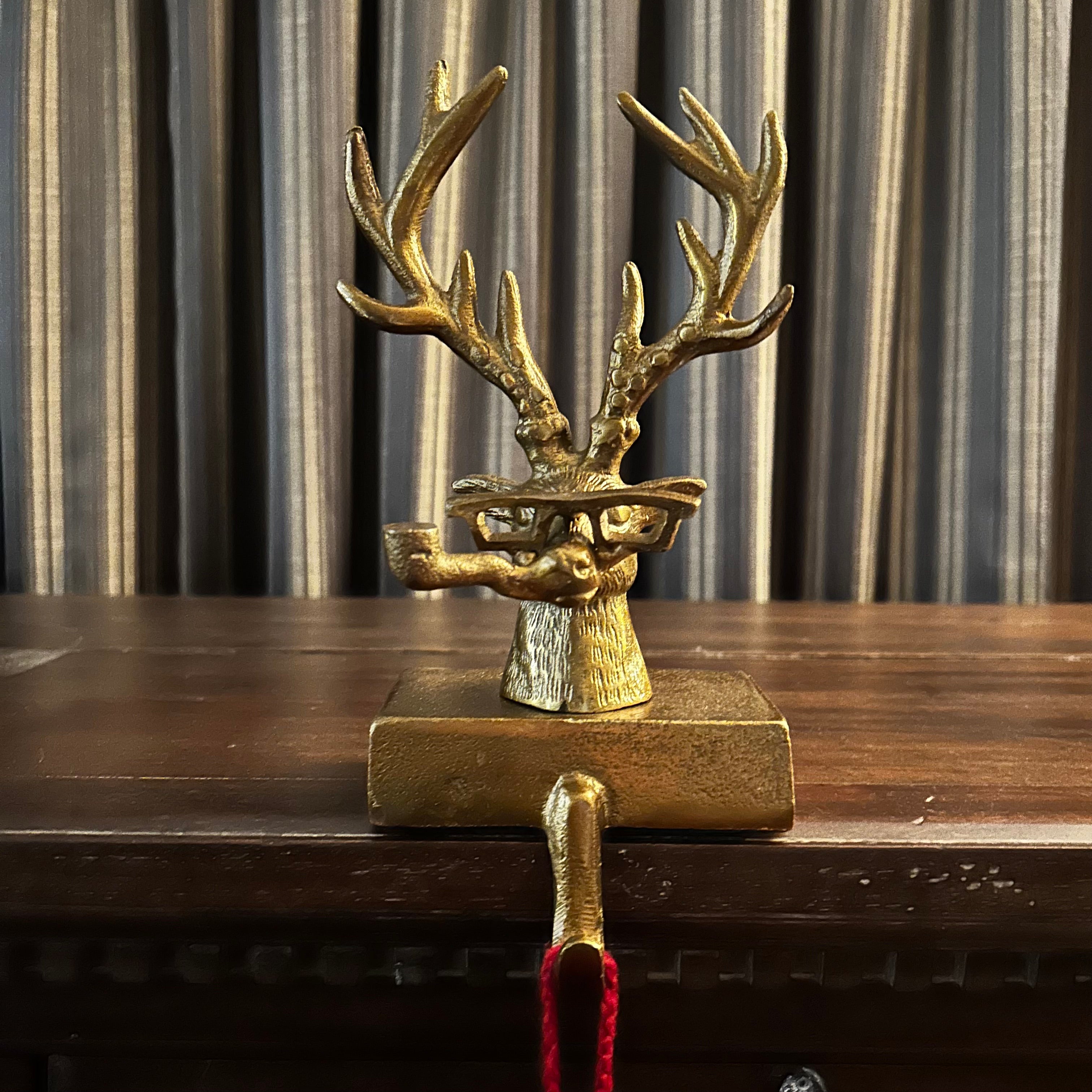 Decorative bronze deer head stocking holder hook on a wooden dresser with striped curtains in the background. The deer is quite a character wearing glasses and smoking a pipe. 