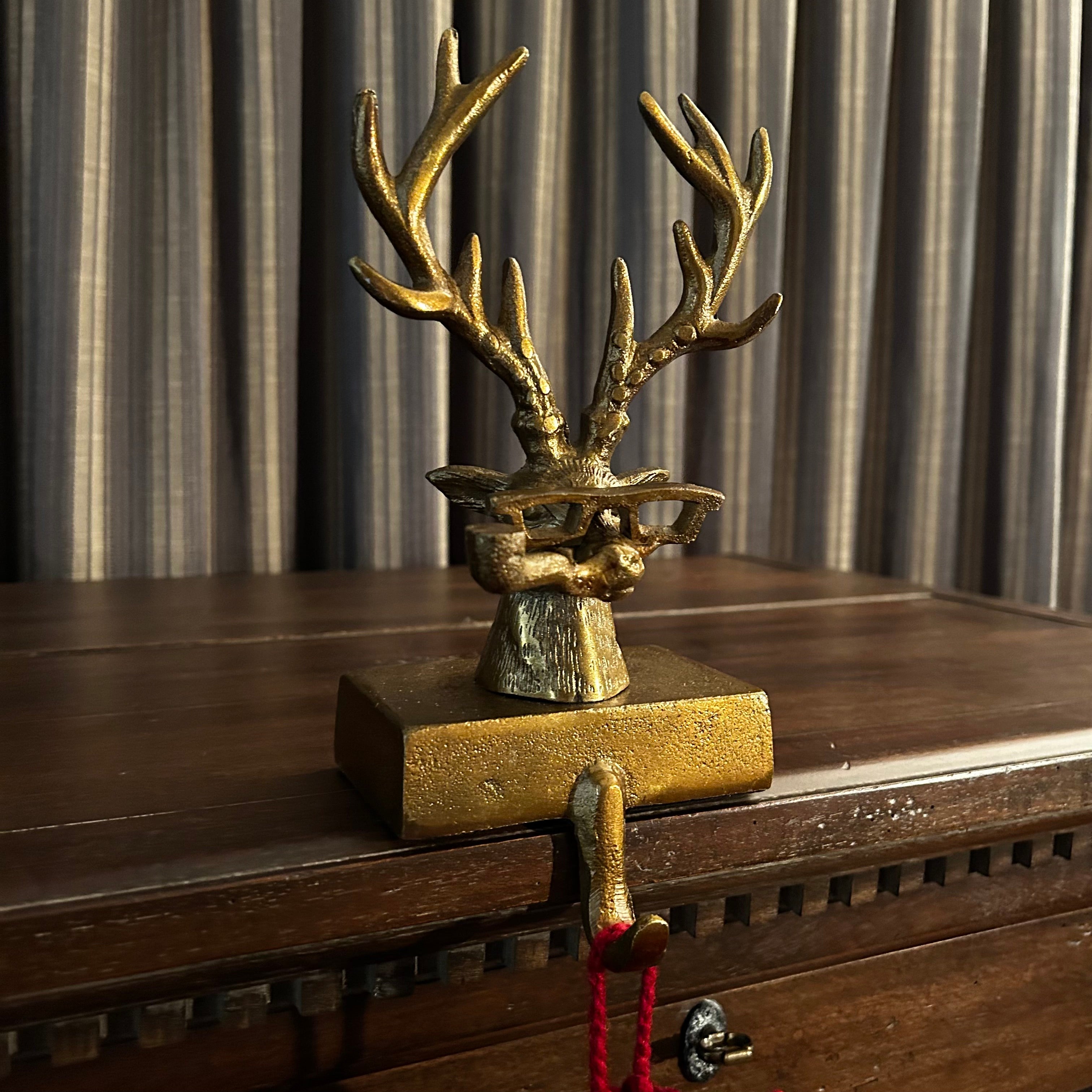 A side view of a decorative bronze deer head stocking holder hook on a wooden dresser with striped curtains in the background. The deer is quite a character wearing glasses and smoking a pipe. 
