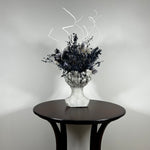This concrete vessel, beautifully sculpted as a woman's serene face, her hair appears to be caught in a soft breeze, with a dried floral arrangement made up navy bupleurum , white curly ting ting, delicate light blue cape flowers, a cluster of three bleached teasels adorns the front left side of her face, and a pure white hydrangea on the right. 