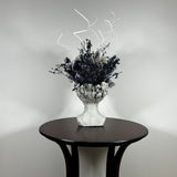 This concrete vessel, beautifully sculpted as a woman's serene face, her hair appears to be caught in a soft breeze, with a dried floral arrangement made up navy bupleurum , white curly ting ting, delicate light blue cape flowers, a cluster of three bleached teasels adorns the front left side of her face, and a pure white hydrangea on the right. 