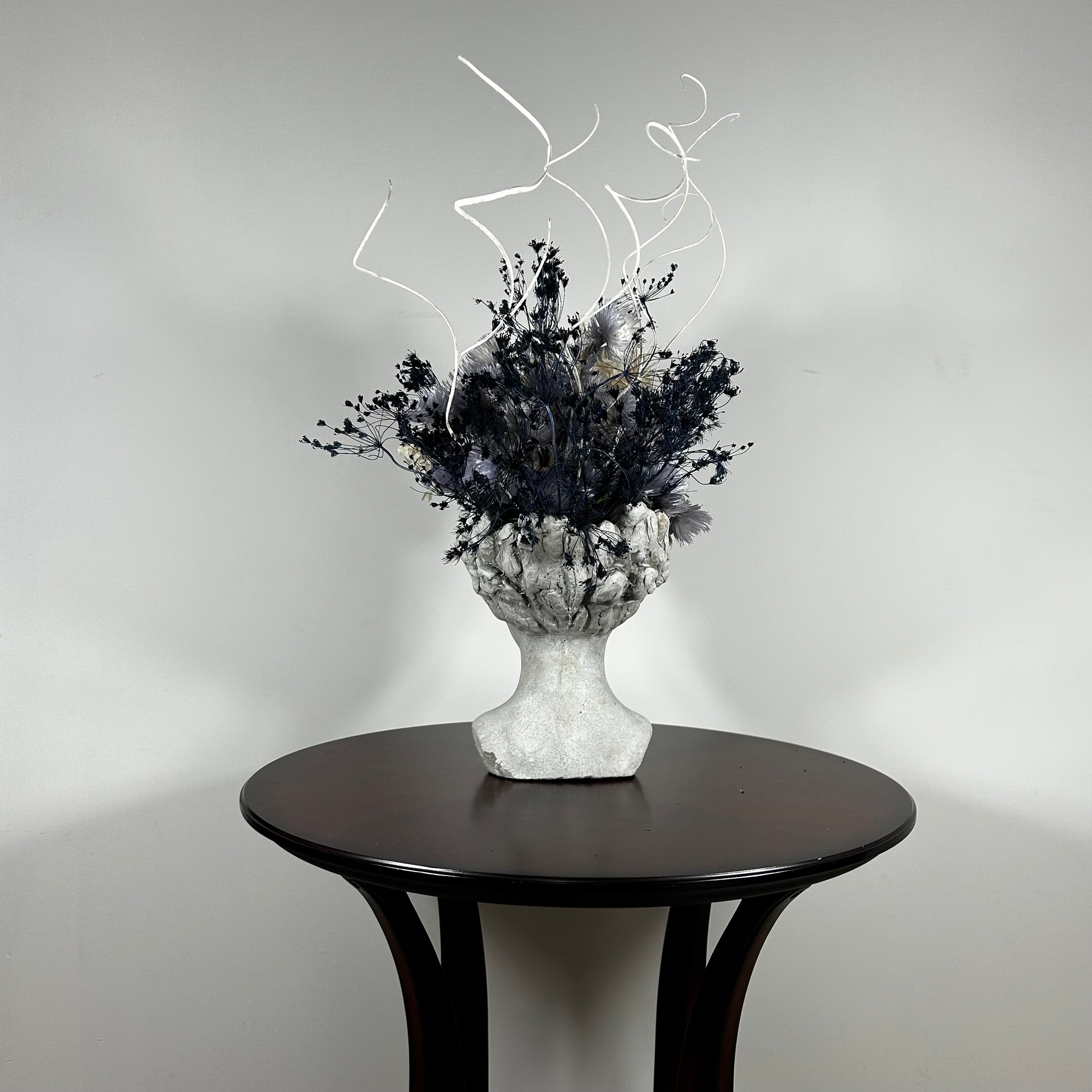 This concrete vessel, beautifully sculpted as a woman's serene face, her hair appears to be caught in a soft breeze, with a dried floral arrangement made up navy bupleurum , white curly ting ting, delicate light blue cape flowers, a cluster of three bleached teasels adorns the front left side of her face, and a pure white hydrangea on the right. 