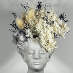 This concrete vessel, beautifully sculpted as a woman's serene face, her hair appears to be caught in a soft breeze, with a dried floral arrangement made up navy bupleurum , white curly ting ting, delicate light blue cape flowers, a cluster of three bleached teasels adorns the front left side of her face, and a pure white hydrangea on the right. 