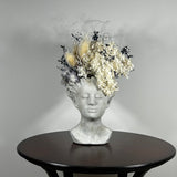 This concrete vessel, beautifully sculpted as a woman's serene face, her hair appears to be caught in a soft breeze, with a dried floral arrangement made up navy bupleurum , white curly ting ting, delicate light blue cape flowers, a cluster of three bleached teasels adorns the front left side of her face, and a pure white hydrangea on the right. 