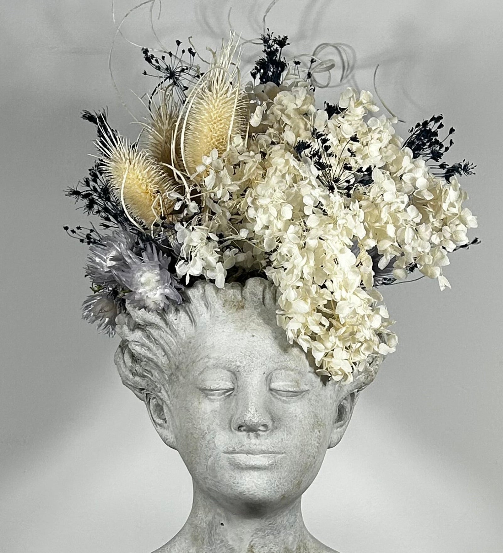 This concrete vessel, beautifully sculpted as a woman's serene face, her hair appears to be caught in a soft breeze, with a dried floral arrangement made up navy bupleurum , white curly ting ting, delicate light blue cape flowers, a cluster of three bleached teasels adorns the front left side of her face, and a pure white hydrangea on the right. 