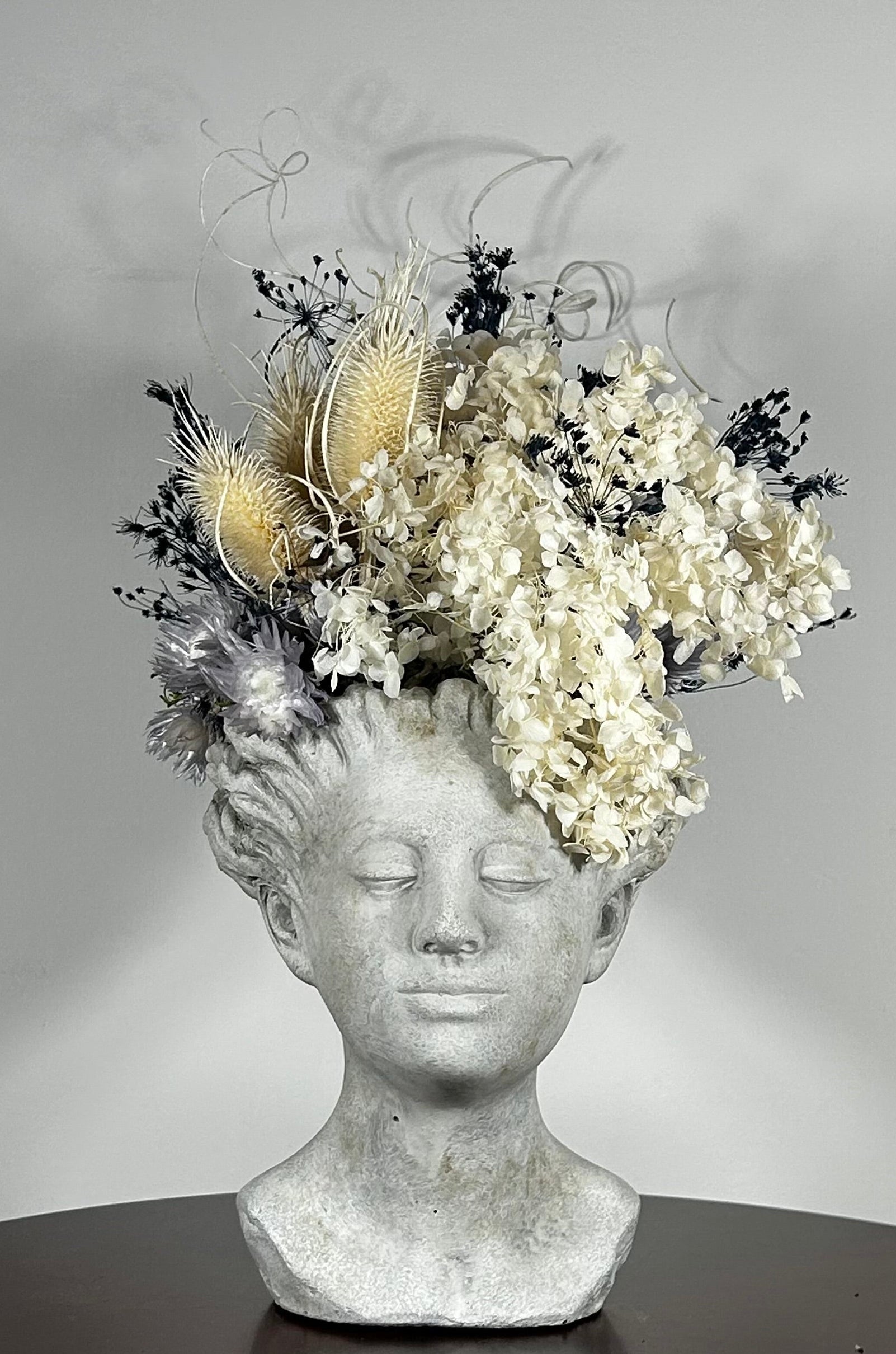 This concrete vessel, beautifully sculpted as a woman's serene face, her hair appears to be caught in a soft breeze, with a dried floral arrangement made up navy bupleurum , white curly ting ting, delicate light blue cape flowers, a cluster of three bleached teasels adorns the front left side of her face, and a pure white hydrangea on the right. 