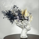 This concrete vessel, beautifully sculpted as a woman's serene face, her hair appears to be caught in a soft breeze, with a dried floral arrangement made up navy bupleurum , white curly ting ting, delicate light blue cape flowers, a cluster of three bleached teasels adorns the front left side of her face, and a pure white hydrangea on the right. 