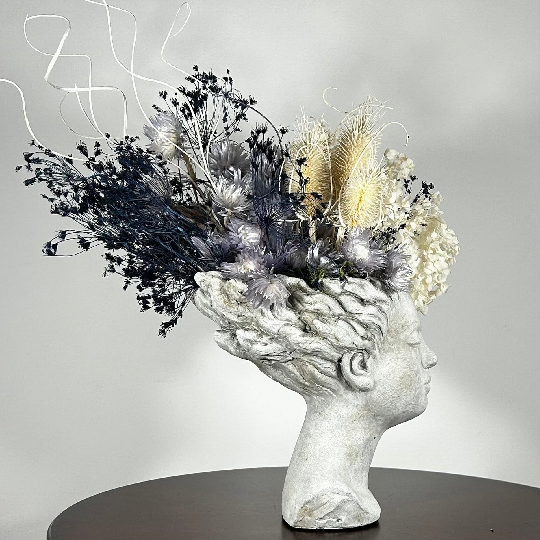 This concrete vessel, beautifully sculpted as a woman's serene face, her hair appears to be caught in a soft breeze, with a dried floral arrangement made up navy bupleurum , white curly ting ting, delicate light blue cape flowers, a cluster of three bleached teasels adorns the front left side of her face, and a pure white hydrangea on the right. 