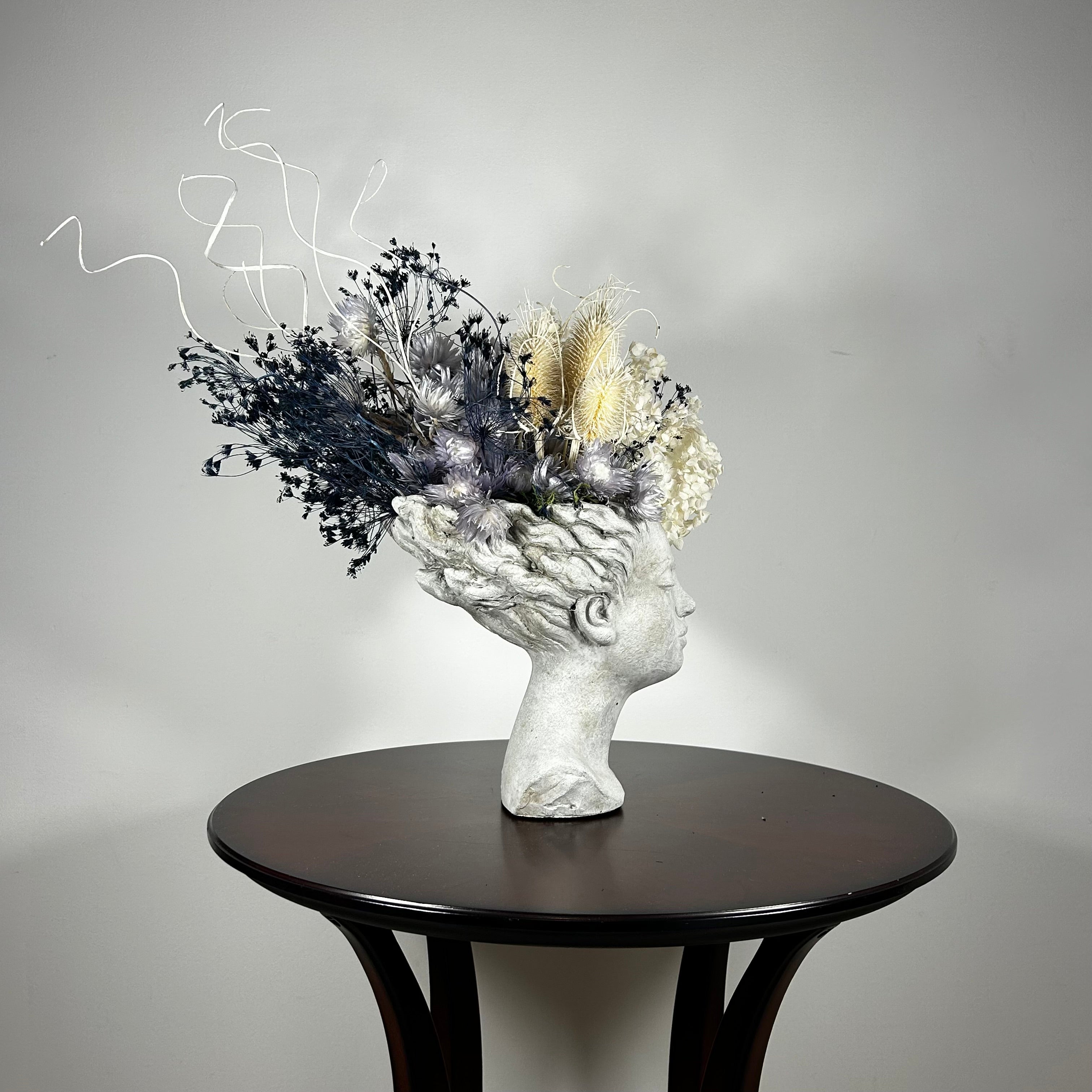 This concrete vessel, beautifully sculpted as a woman's serene face, her hair appears to be caught in a soft breeze, with a dried floral arrangement made up navy bupleurum , white curly ting ting, delicate light blue cape flowers, a cluster of three bleached teasels adorns the front left side of her face, and a pure white hydrangea on the right. 
