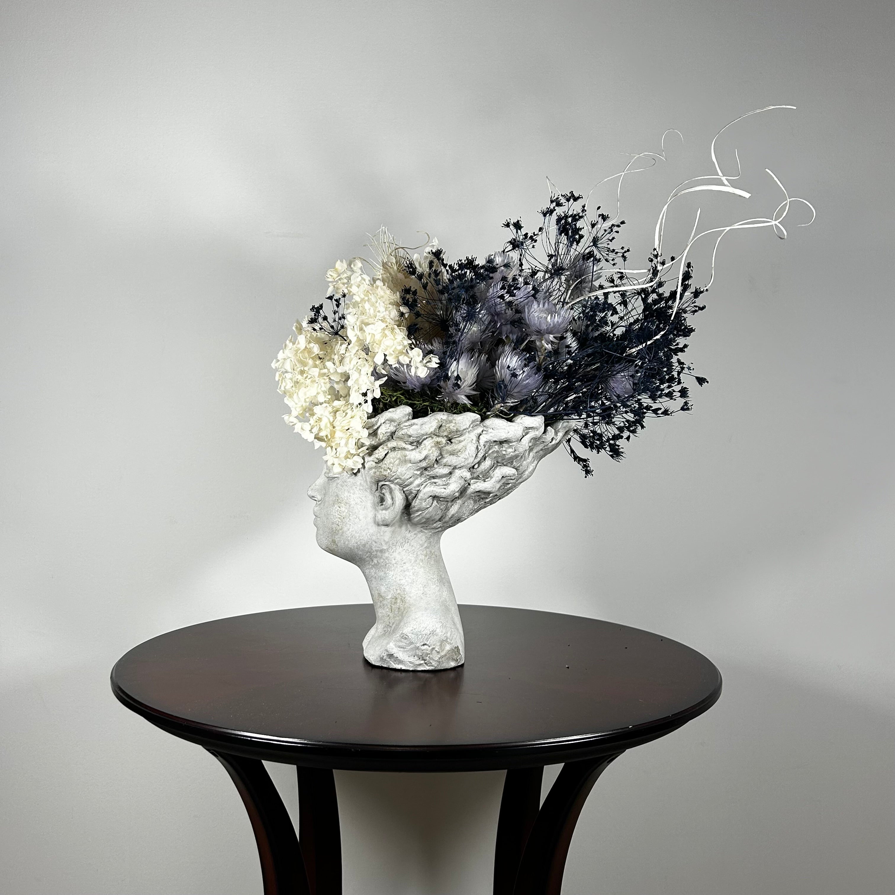 This concrete vessel, beautifully sculpted as a woman's serene face, her hair appears to be caught in a soft breeze, with a dried floral arrangement made up navy bupleurum , white curly ting ting, delicate light blue cape flowers, a cluster of three bleached teasels adorns the front left side of her face, and a pure white hydrangea on the right. 