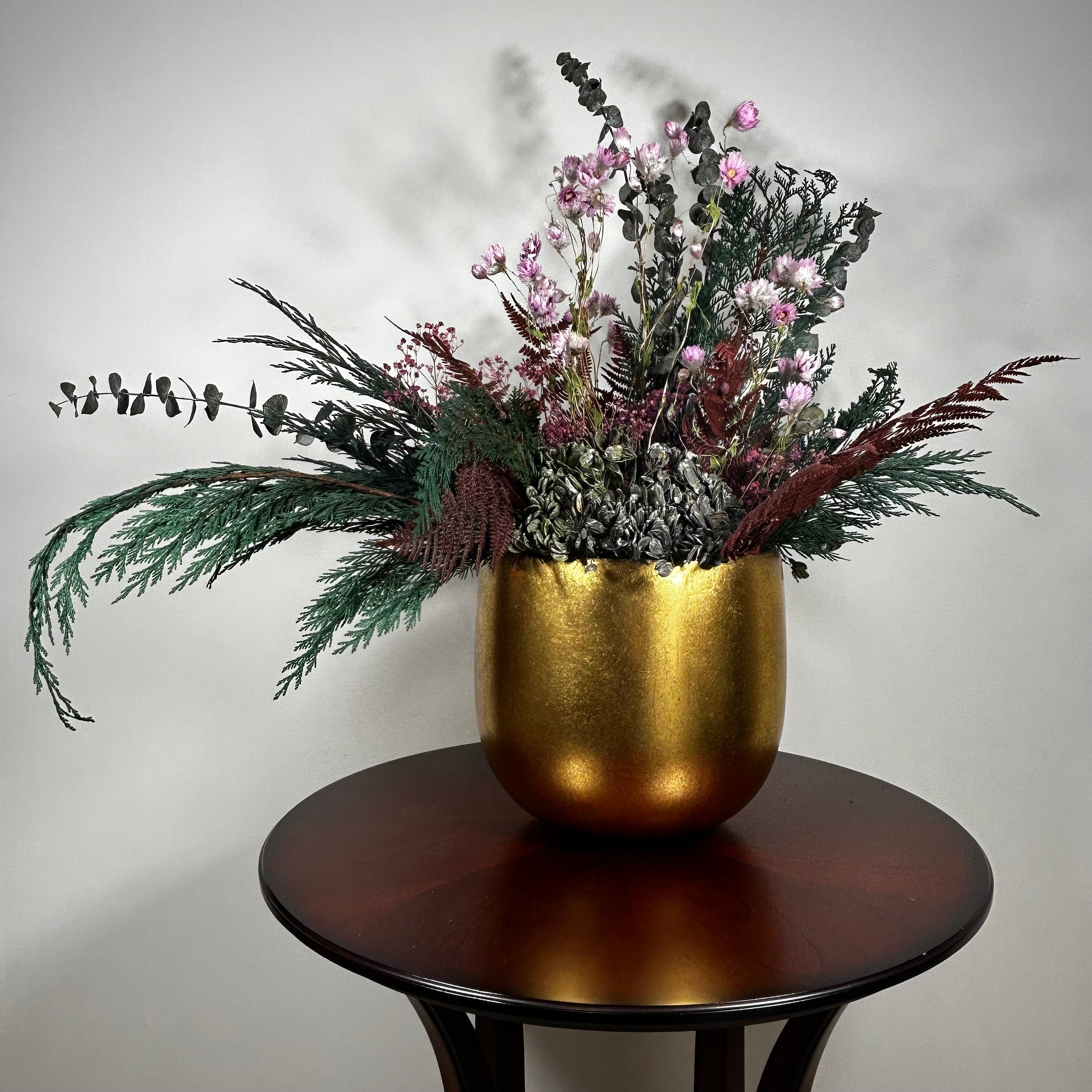 Matte gold cylinder pot holds greens of preserved Cedar and the beautiful deep red tones of preserved Bracken. Contrast is achieved through the icy elegance of preserved blue frosted Eucalyptus and the unique, ethereal fluff of dried frosted Hogweed. Delicate Acroclinium (paper daisies) in natural pink float above the bulk of the arrangement, complemented by the rich colour of Gypsophila (baby's breath) dyed deep rose.