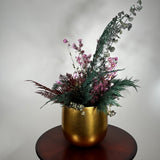 Matte gold cylinder pot holds greens of preserved Cedar and the beautiful deep red tones of preserved Bracken. Contrast is achieved through the icy elegance of preserved blue frosted Eucalyptus and the unique, ethereal fluff of dried frosted Hogweed. Delicate Acroclinium (paper daisies) in natural pink float above the bulk of the arrangement, complemented by the rich colour of Gypsophila (baby's breath) dyed deep rose.