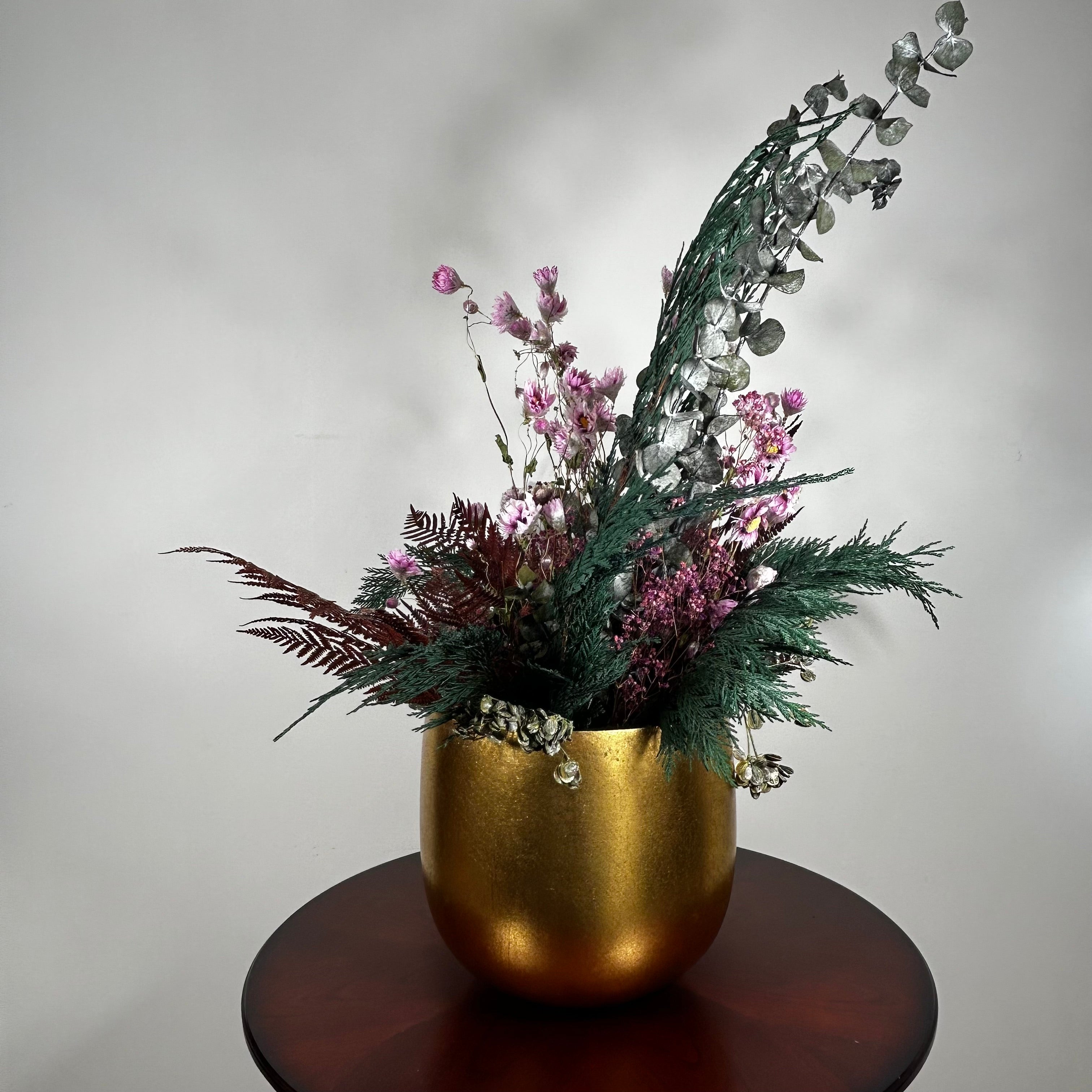Matte gold cylinder pot holds greens of preserved Cedar and the beautiful deep red tones of preserved Bracken. Contrast is achieved through the icy elegance of preserved blue frosted Eucalyptus and the unique, ethereal fluff of dried frosted Hogweed. Delicate Acroclinium (paper daisies) in natural pink float above the bulk of the arrangement, complemented by the rich colour of Gypsophila (baby's breath) dyed deep rose.