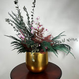 Matte gold cylinder pot holds greens of preserved Cedar and the beautiful deep red tones of preserved Bracken. Contrast is achieved through the icy elegance of preserved blue frosted Eucalyptus and the unique, ethereal fluff of dried frosted Hogweed. Delicate Acroclinium (paper daisies) in natural pink float above the bulk of the arrangement, complemented by the rich colour of Gypsophila (baby's breath) dyed deep rose.
