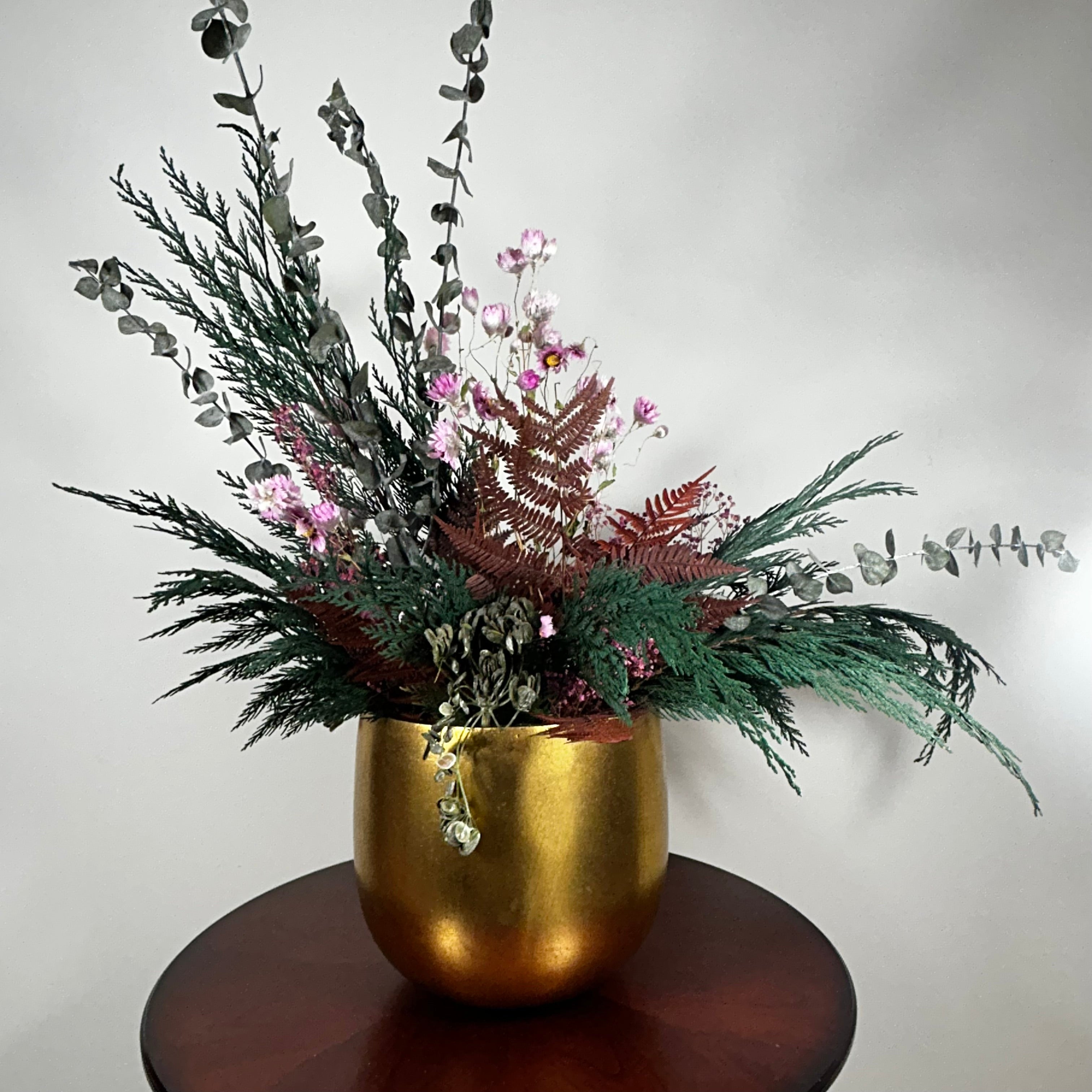 Matte gold cylinder pot holds greens of preserved Cedar and the beautiful deep red tones of preserved Bracken. Contrast is achieved through the icy elegance of preserved blue frosted Eucalyptus and the unique, ethereal fluff of dried frosted Hogweed. Delicate Acroclinium (paper daisies) in natural pink float above the bulk of the arrangement, complemented by the rich colour of Gypsophila (baby's breath) dyed deep rose.