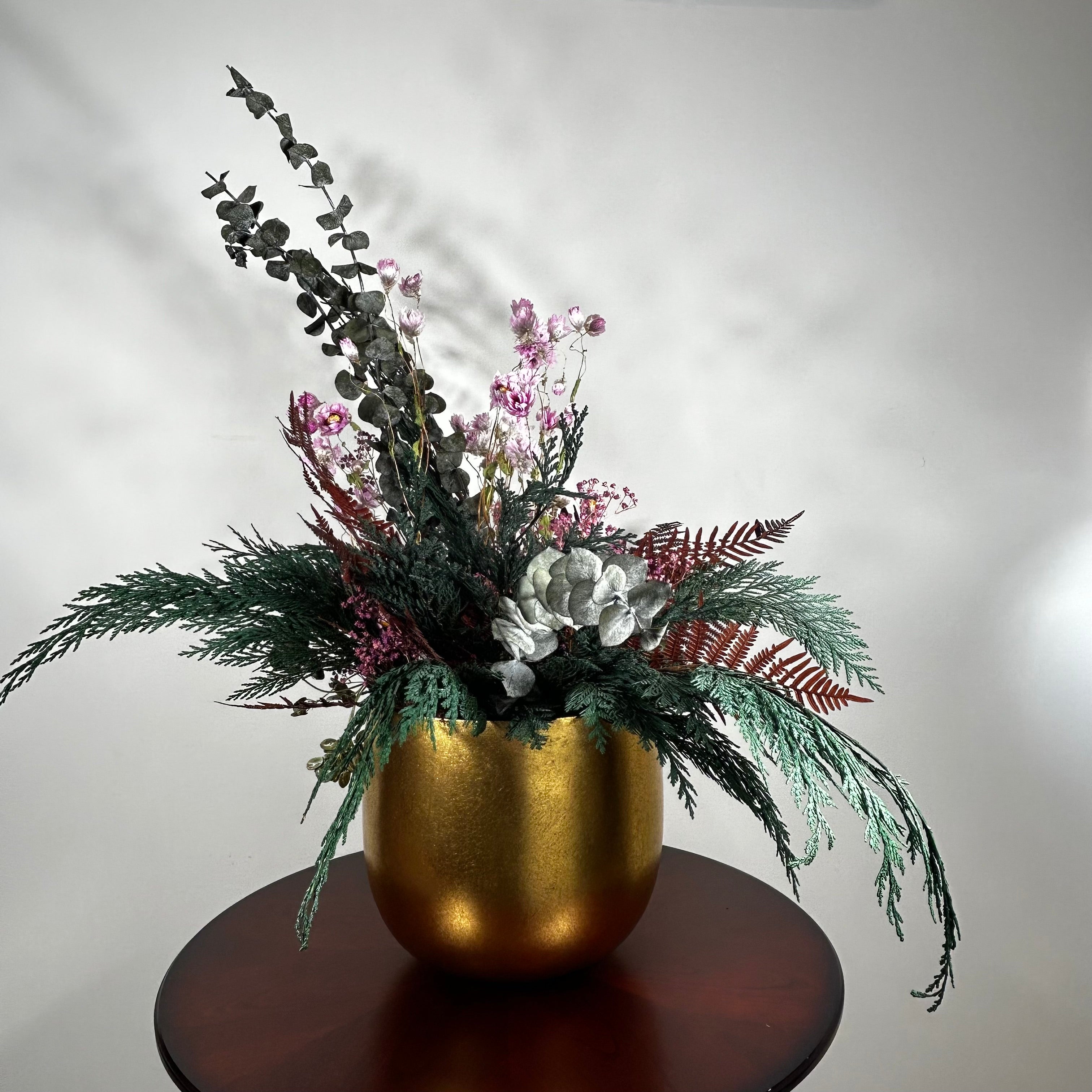 Matte gold cylinder pot holds greens of preserved Cedar and the beautiful deep red tones of preserved Bracken. Contrast is achieved through the icy elegance of preserved blue frosted Eucalyptus and the unique, ethereal fluff of dried frosted Hogweed. Delicate Acroclinium (paper daisies) in natural pink float above the bulk of the arrangement, complemented by the rich colour of Gypsophila (baby's breath) dyed deep rose.