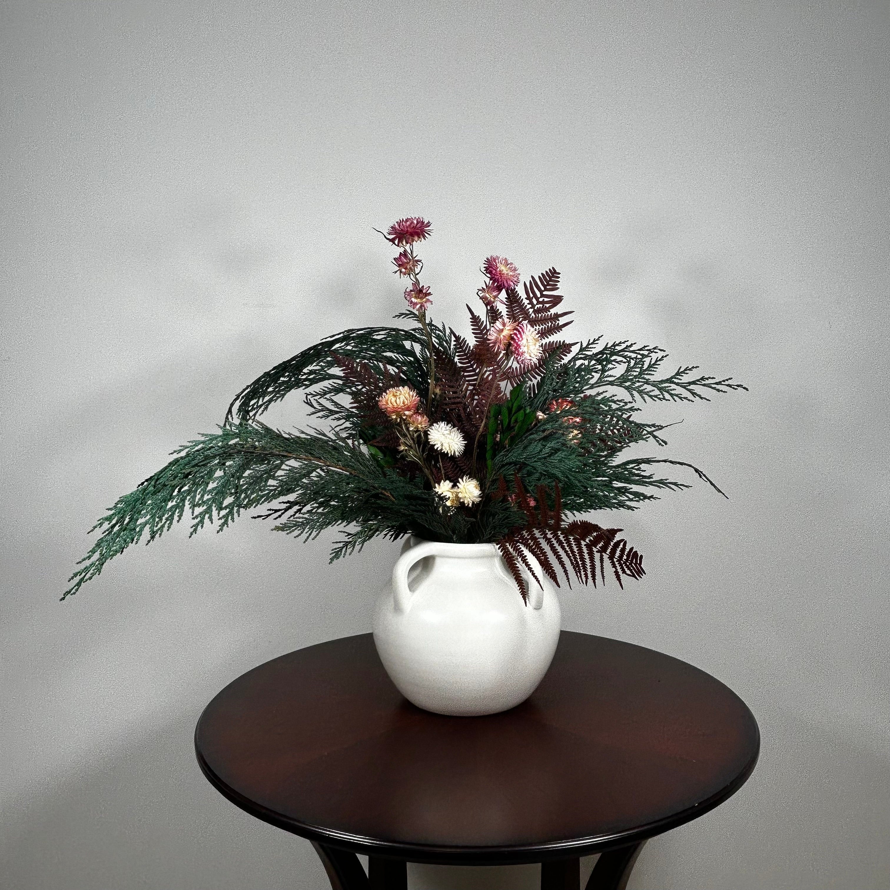 Dried floral arrangement in a matte white ceramic vase filled with richly textured preserved greens, including aromatic preserved Cedar and dense preserved Boxwood. This traditional base provides a stunning contrast to the striking accents: deep preserved Burgundy Bracken adds dramatic colour, while natural pink Helichrysums (strawflowers) provide a gentle floral lift.