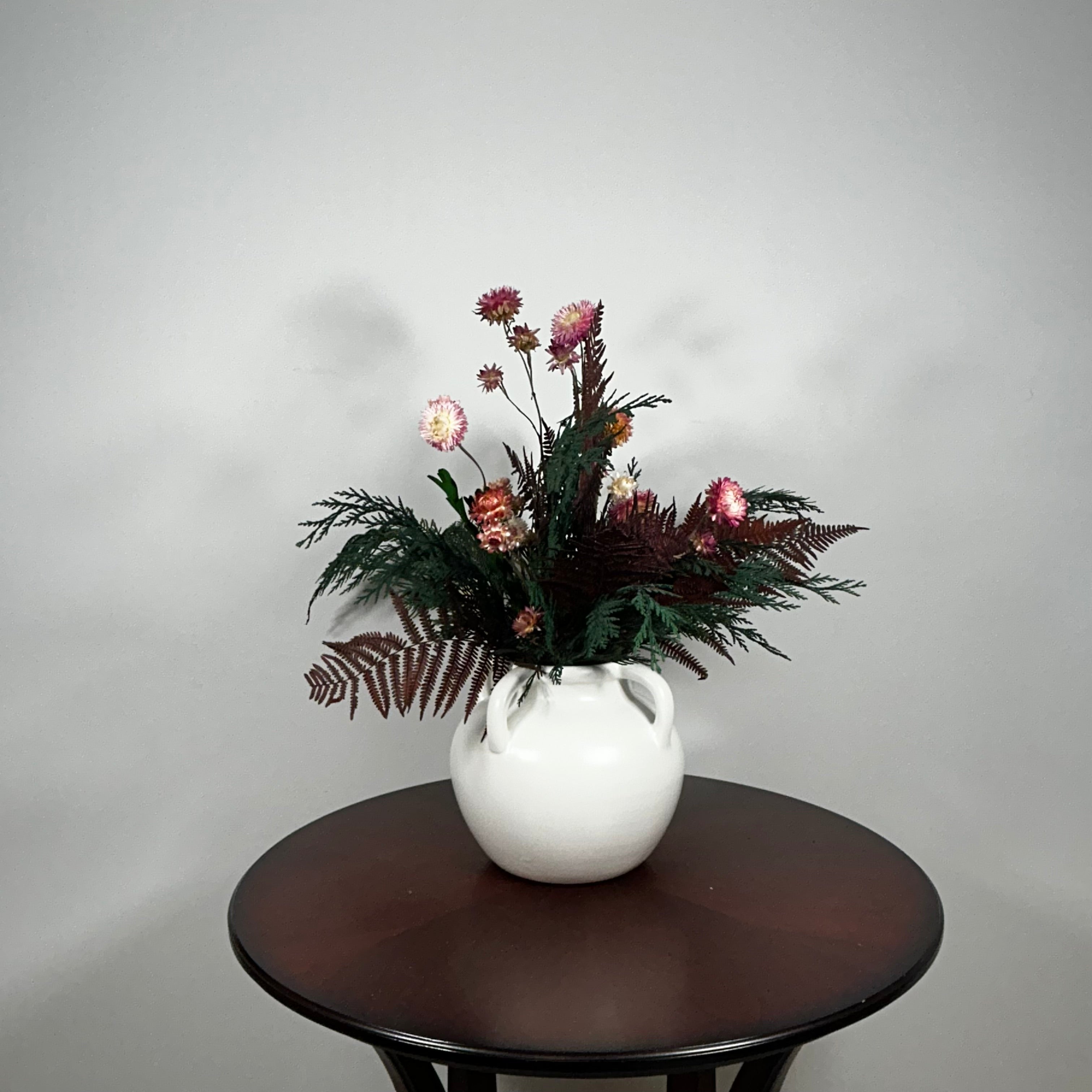 Dried floral arrangement in a matte white ceramic vase filled with richly textured preserved greens, including aromatic preserved Cedar and dense preserved Boxwood. This traditional base provides a stunning contrast to the striking accents: deep preserved Burgundy Bracken adds dramatic colour, while natural pink Helichrysums (strawflowers) provide a gentle floral lift.