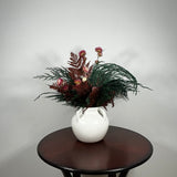Dried floral arrangement in a matte white ceramic vase filled with richly textured preserved greens, including aromatic preserved Cedar and dense preserved Boxwood. This traditional base provides a stunning contrast to the striking accents: deep preserved Burgundy Bracken adds dramatic colour, while natural pink Helichrysums (strawflowers) provide a gentle floral lift.