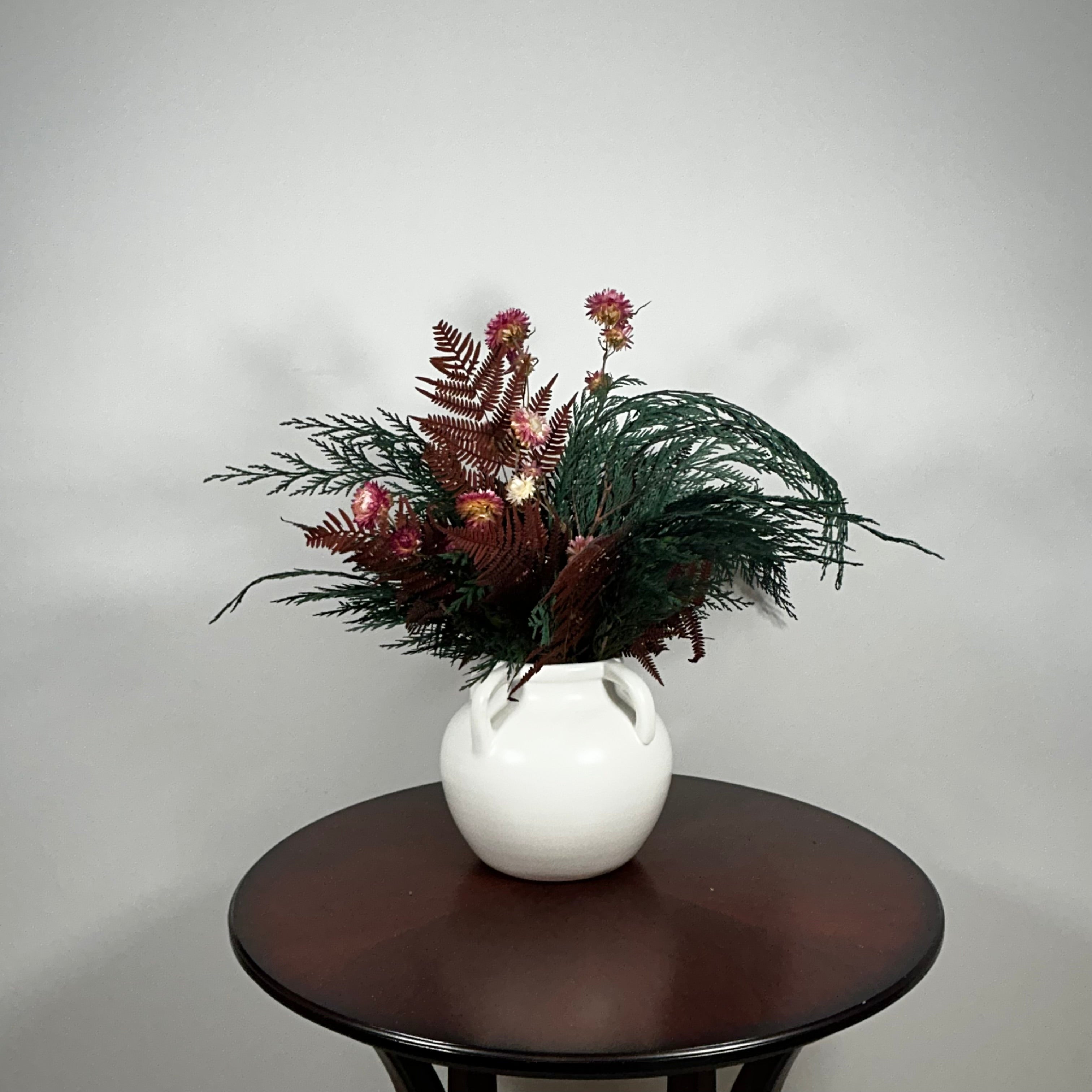Dried floral arrangement in a matte white ceramic vase filled with richly textured preserved greens, including aromatic preserved Cedar and dense preserved Boxwood. This traditional base provides a stunning contrast to the striking accents: deep preserved Burgundy Bracken adds dramatic colour, while natural pink Helichrysums (strawflowers) provide a gentle floral lift.