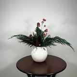 Dried floral arrangement in a matte white ceramic vase filled with richly textured preserved greens, including aromatic preserved Cedar and dense preserved Boxwood. This traditional base provides a stunning contrast to the striking accents: deep preserved Burgundy Bracken adds dramatic colour, while natural pink Helichrysums (strawflowers) provide a gentle floral lift.