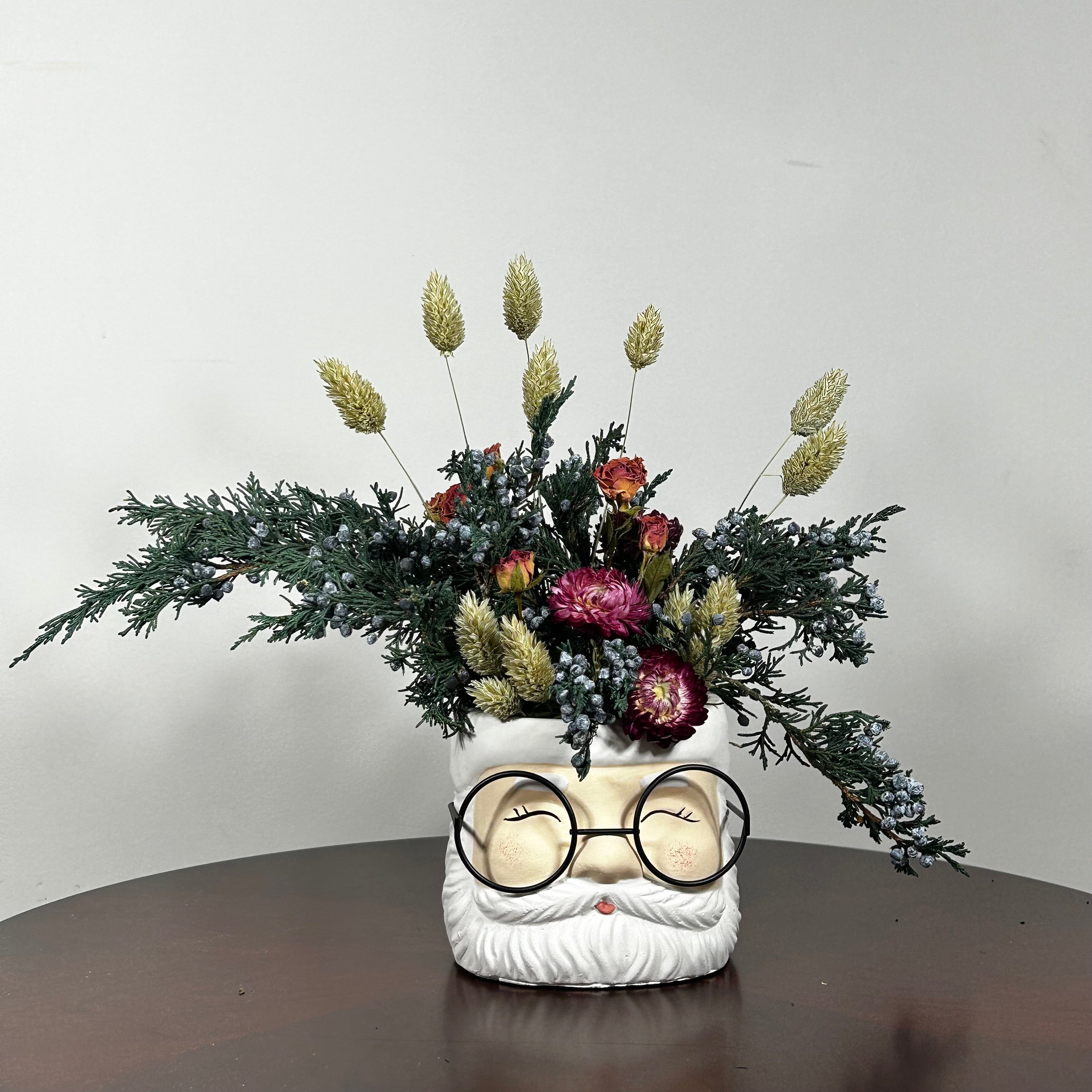 A dried floral arrangement in a Santa face pot featuring preserved Christmas greenery from fragrant Juniper branches with their distinctive blue berries. This is beautifully contrasted with the soft, elegant charm of pink rose clusters and the bold red color of deep red strawflowers. The entire composition is softened by the airy, feathery texture of natural Phalaris (Canary Grass), which floats above the bulk of the arrangement.
