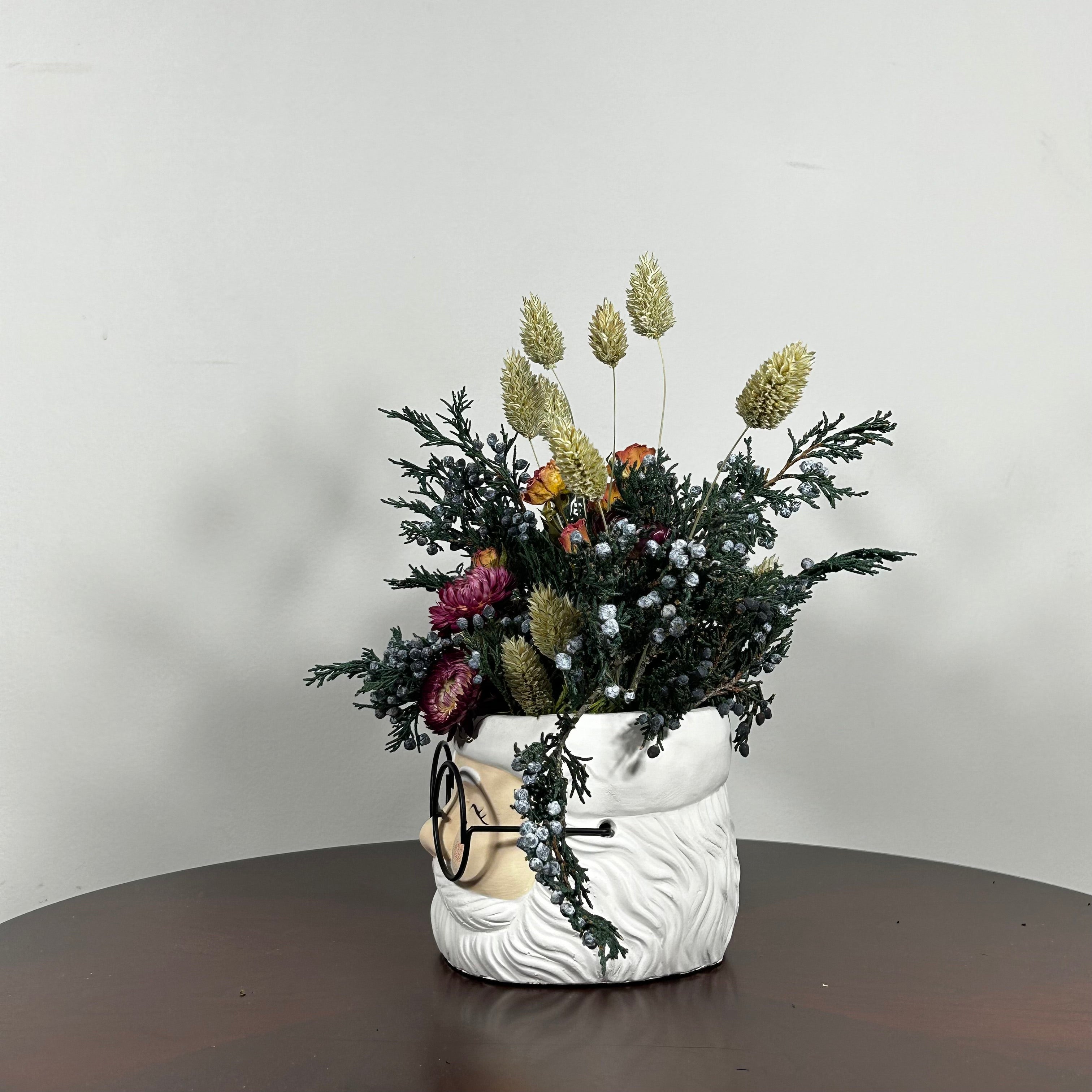 Side view of a dried floral arrangement in a Santa face pot featuring preserved Christmas greenery from fragrant Juniper branches with their distinctive blue berries. This is beautifully contrasted with the soft, elegant charm of pink rose clusters and the bold red color of deep red strawflowers. The entire composition is softened by the airy, feathery texture of natural Phalaris (Canary Grass), which floats above the bulk of the arrangement.
