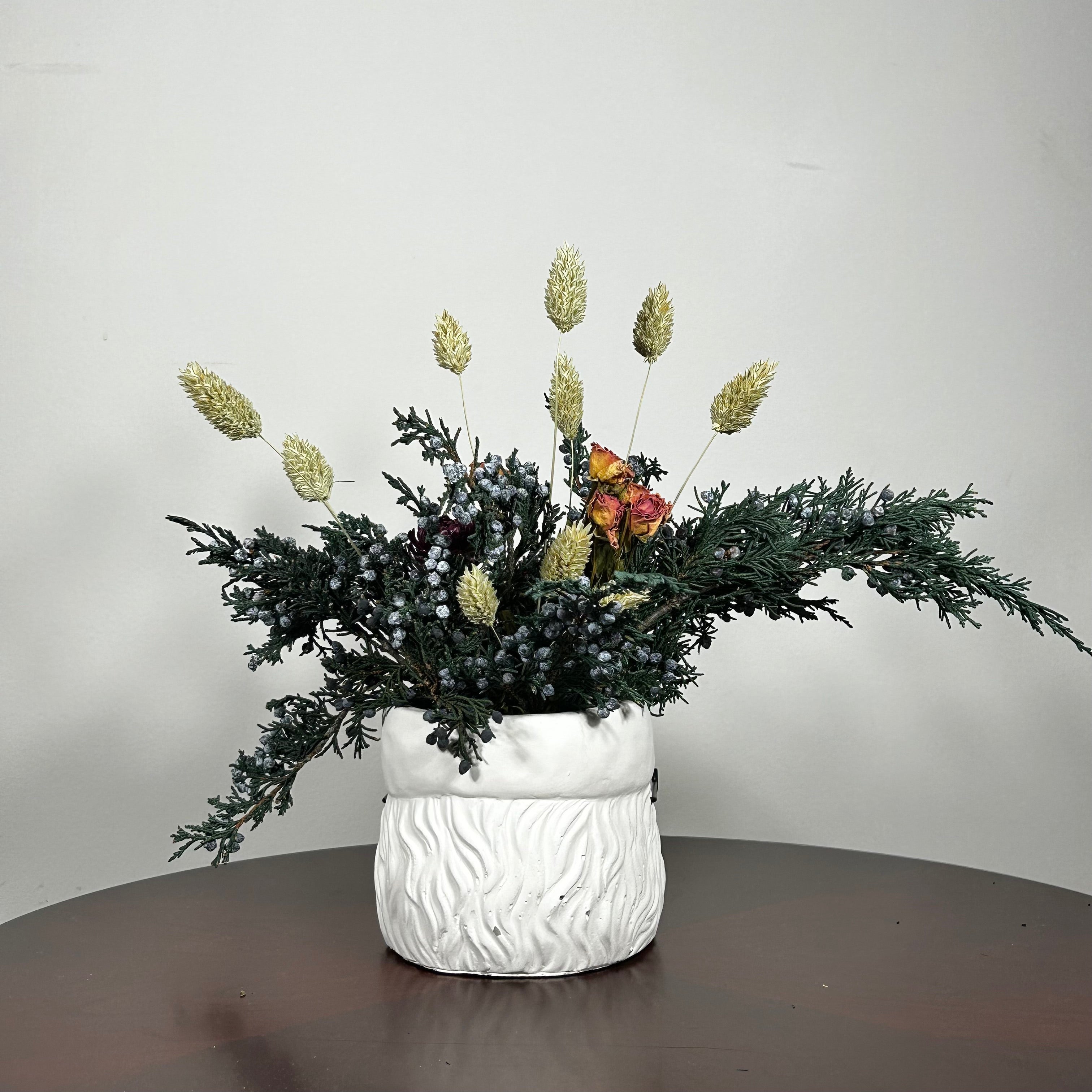 Back view of a dried floral arrangement in a Santa face pot featuring preserved Christmas greenery from fragrant Juniper branches with their distinctive blue berries. This is beautifully contrasted with the soft, elegant charm of pink rose clusters and the bold red color of deep red strawflowers. The entire composition is softened by the airy, feathery texture of natural Phalaris (Canary Grass), which floats above the bulk of the arrangement.

