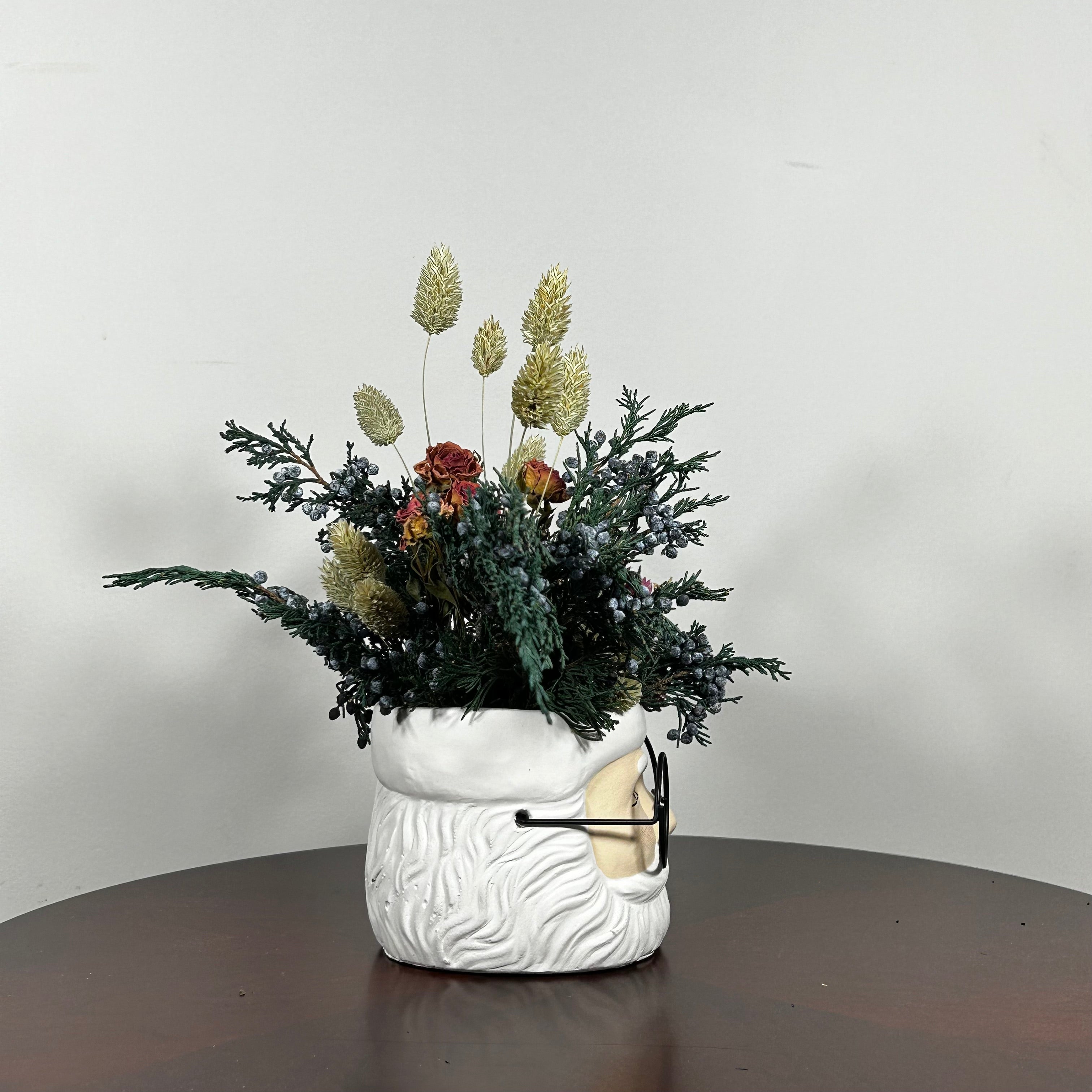 Side view of a dried floral arrangement in a Santa face pot featuring preserved Christmas greenery from fragrant Juniper branches with their distinctive blue berries. This is beautifully contrasted with the soft, elegant charm of pink rose clusters and the bold red color of deep red strawflowers. The entire composition is softened by the airy, feathery texture of natural Phalaris (Canary Grass), which floats above the bulk of the arrangement.