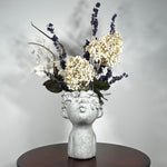 A dried flower arrangement in a concrete vessel resembling a face with puckered lips waiting to be kissed. The florals include lush purple Larkspur reaching skyward, complemented by the subtle charm of pale lime Hydrangea and the bright purity of white Luminary. Adding a touch of rustic artistry, three brown curly cocoa sticks create dynamic visual interest. The entire composition is gracefully grounded by verdant preserved Salal leaves, completing this ethereal blend of natural beauty and playful artistry.
