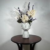A dried flower arrangement in a concrete vessel resembling a face with puckered lips waiting to be kissed. The florals include lush purple Larkspur reaching skyward, complemented by the subtle charm of pale lime Hydrangea and the bright purity of white Luminary. Adding a touch of rustic artistry, three brown curly cocoa sticks create dynamic visual interest. The entire composition is gracefully grounded by verdant preserved Salal leaves, completing this ethereal blend of natural beauty and playful artistry.