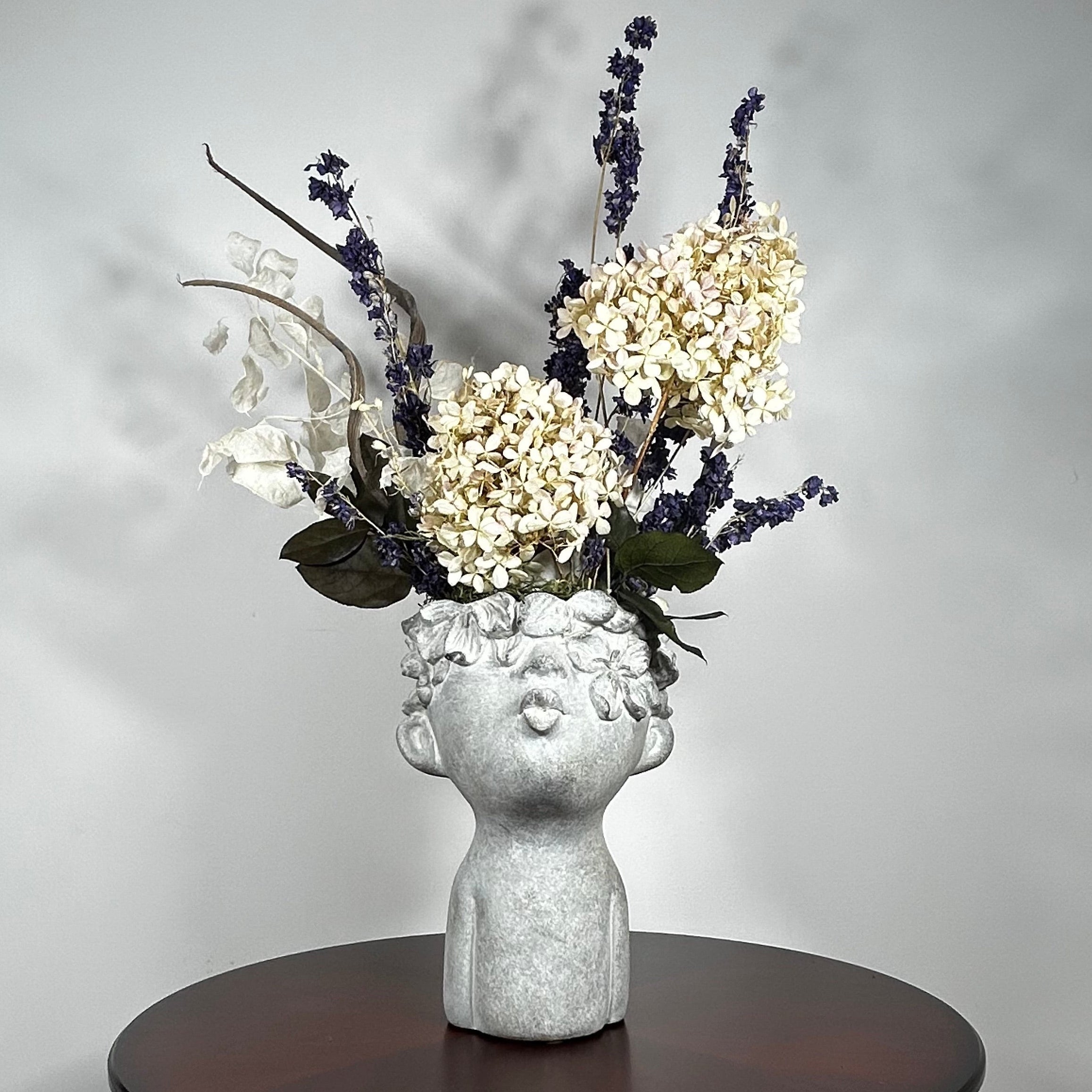 A dried flower arrangement in a concrete vessel resembling a face with puckered lips waiting to be kissed. The florals include lush purple Larkspur reaching skyward, complemented by the subtle charm of pale lime Hydrangea and the bright purity of white Luminary. Adding a touch of rustic artistry, three brown curly cocoa sticks create dynamic visual interest. The entire composition is gracefully grounded by verdant preserved Salal leaves, completing this ethereal blend of natural beauty and playful artistry.
