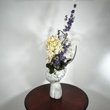 A dried flower arrangement in a concrete vessel resembling a face with puckered lips waiting to be kissed. The florals include lush purple Larkspur reaching skyward, complemented by the subtle charm of pale lime Hydrangea and the bright purity of white Luminary. Adding a touch of rustic artistry, three brown curly cocoa sticks create dynamic visual interest. The entire composition is gracefully grounded by verdant preserved Salal leaves, completing this ethereal blend of natural beauty and playful artistry.