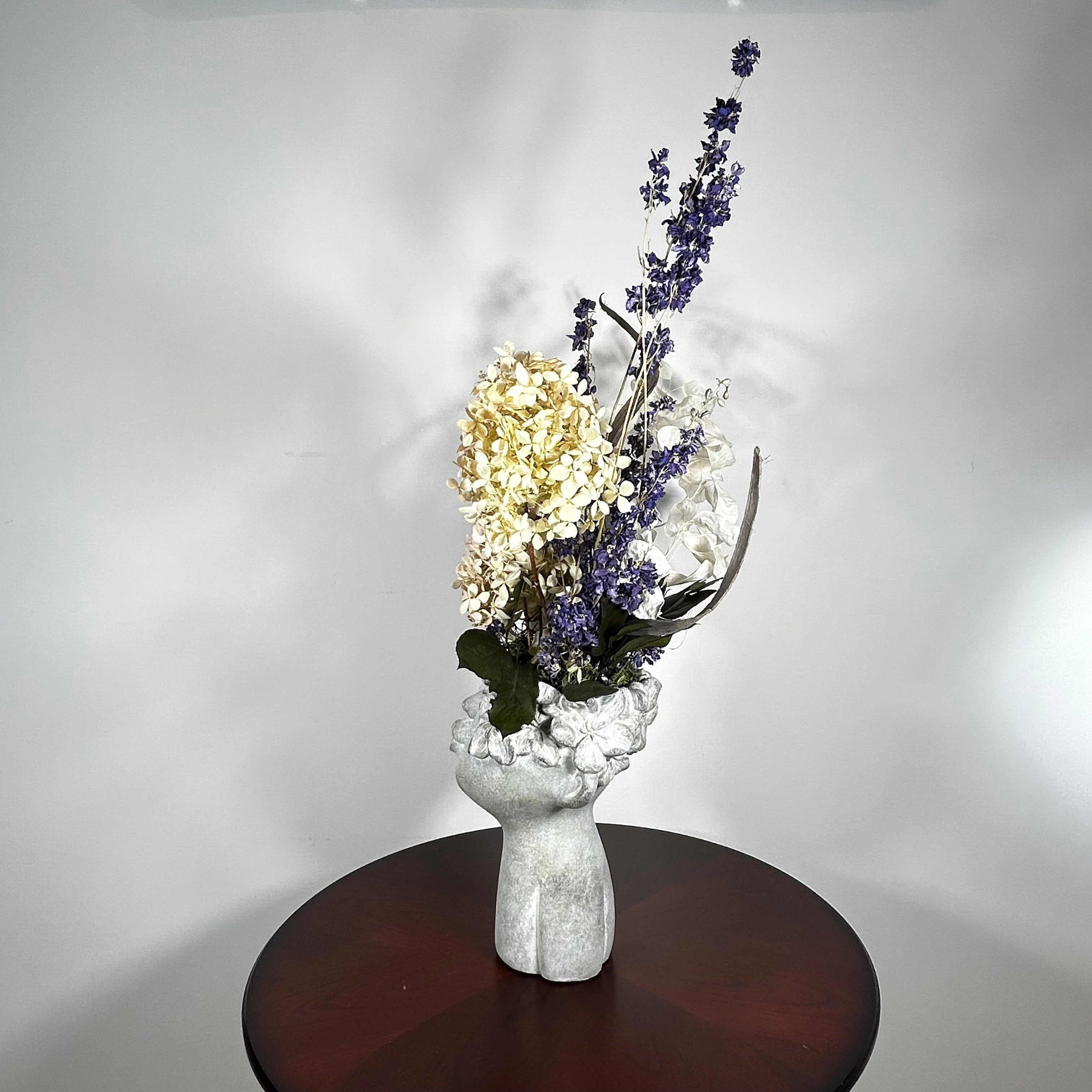 A dried flower arrangement in a concrete vessel resembling a face with puckered lips waiting to be kissed. The florals include lush purple Larkspur reaching skyward, complemented by the subtle charm of pale lime Hydrangea and the bright purity of white Luminary. Adding a touch of rustic artistry, three brown curly cocoa sticks create dynamic visual interest. The entire composition is gracefully grounded by verdant preserved Salal leaves, completing this ethereal blend of natural beauty and playful artistry.