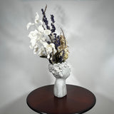 A dried flower arrangement in a concrete vessel resembling a face with puckered lips waiting to be kissed. The florals include lush purple Larkspur reaching skyward, complemented by the subtle charm of pale lime Hydrangea and the bright purity of white Luminary. Adding a touch of rustic artistry, three brown curly cocoa sticks create dynamic visual interest. The entire composition is gracefully grounded by verdant preserved Salal leaves, completing this ethereal blend of natural beauty and playful artistry.