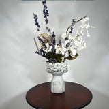 A dried flower arrangement in a concrete vessel resembling a face with puckered lips waiting to be kissed. The florals include lush purple Larkspur reaching skyward, complemented by the subtle charm of pale lime Hydrangea and the bright purity of white Luminary. Adding a touch of rustic artistry, three brown curly cocoa sticks create dynamic visual interest. The entire composition is gracefully grounded by verdant preserved Salal leaves, completing this ethereal blend of natural beauty and playful artistry.