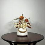 Dried flower arrangement with slender stems of natural green willow eucalyptus reach out from one side, complemented on the opposite side by the soft, bleached plumes of Calamgrostis canadensis. Ethereal Lunaria in pristine white adds a delicate, luminous quality. The featured floral is a cluster of five Dahlia stems, their petals a beautiful rust tone that gracefully fades into a vibrant golden heart, a perfect echo of the setting sun.