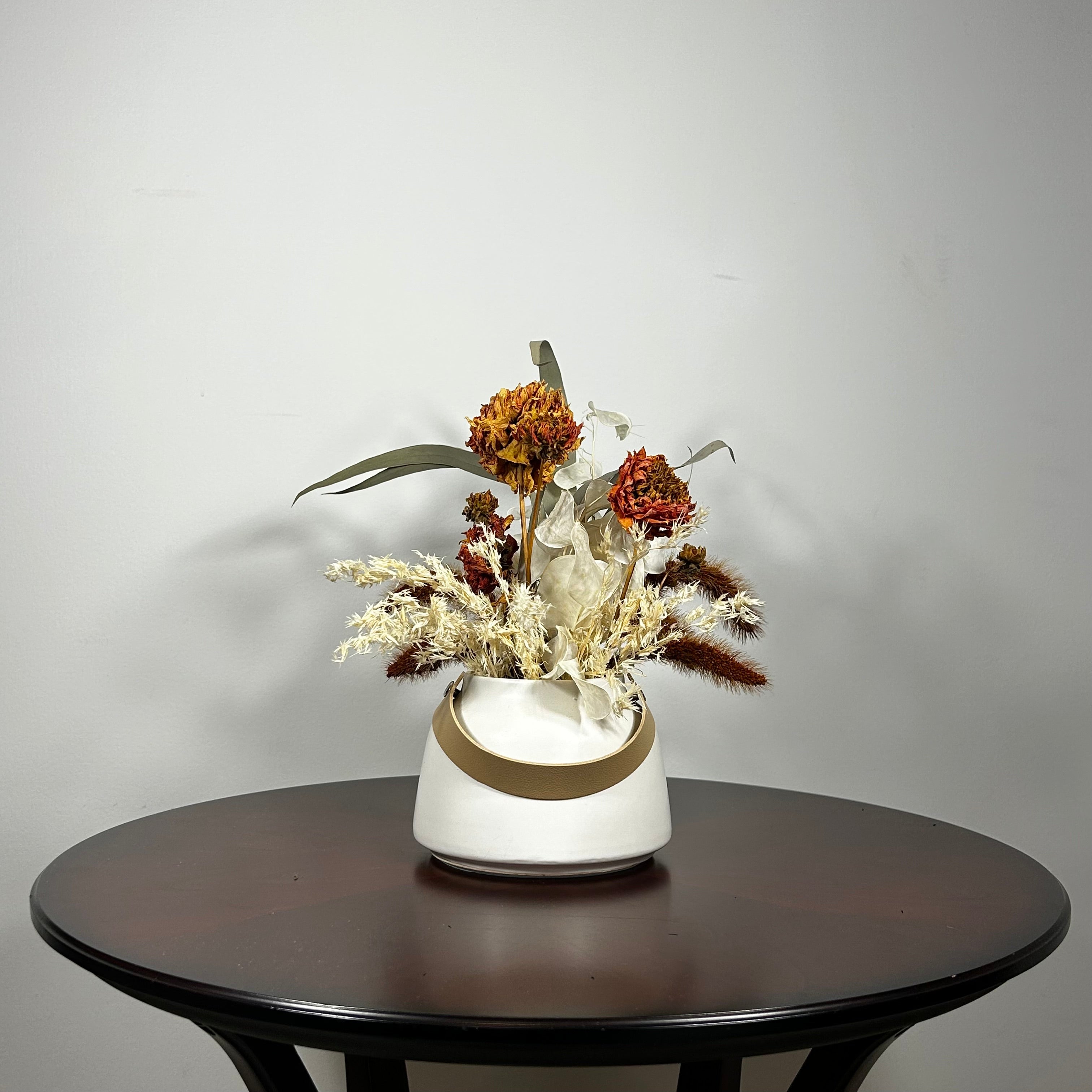 Dried flower arrangement with slender stems of natural green willow eucalyptus reach out from one side, complemented on the opposite side by the soft, bleached plumes of Calamgrostis canadensis. Ethereal Lunaria in pristine white adds a delicate, luminous quality. The featured floral is a cluster of five Dahlia stems, their petals a beautiful rust tone that gracefully fades into a vibrant golden heart, a perfect echo of the setting sun.