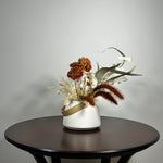 Dried flower arrangement with slender stems of natural green willow eucalyptus reach out from one side, complemented on the opposite side by the soft, bleached plumes of Calamgrostis canadensis. Ethereal Lunaria in pristine white adds a delicate, luminous quality. The featured floral is a cluster of five Dahlia stems, their petals a beautiful rust tone that gracefully fades into a vibrant golden heart, a perfect echo of the setting sun.