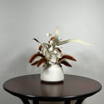 Dried flower arrangement with slender stems of natural green willow eucalyptus reach out from one side, complemented on the opposite side by the soft, bleached plumes of Calamgrostis canadensis. Ethereal Lunaria in pristine white adds a delicate, luminous quality. The featured floral is a cluster of five Dahlia stems, their petals a beautiful rust tone that gracefully fades into a vibrant golden heart, a perfect echo of the setting sun.