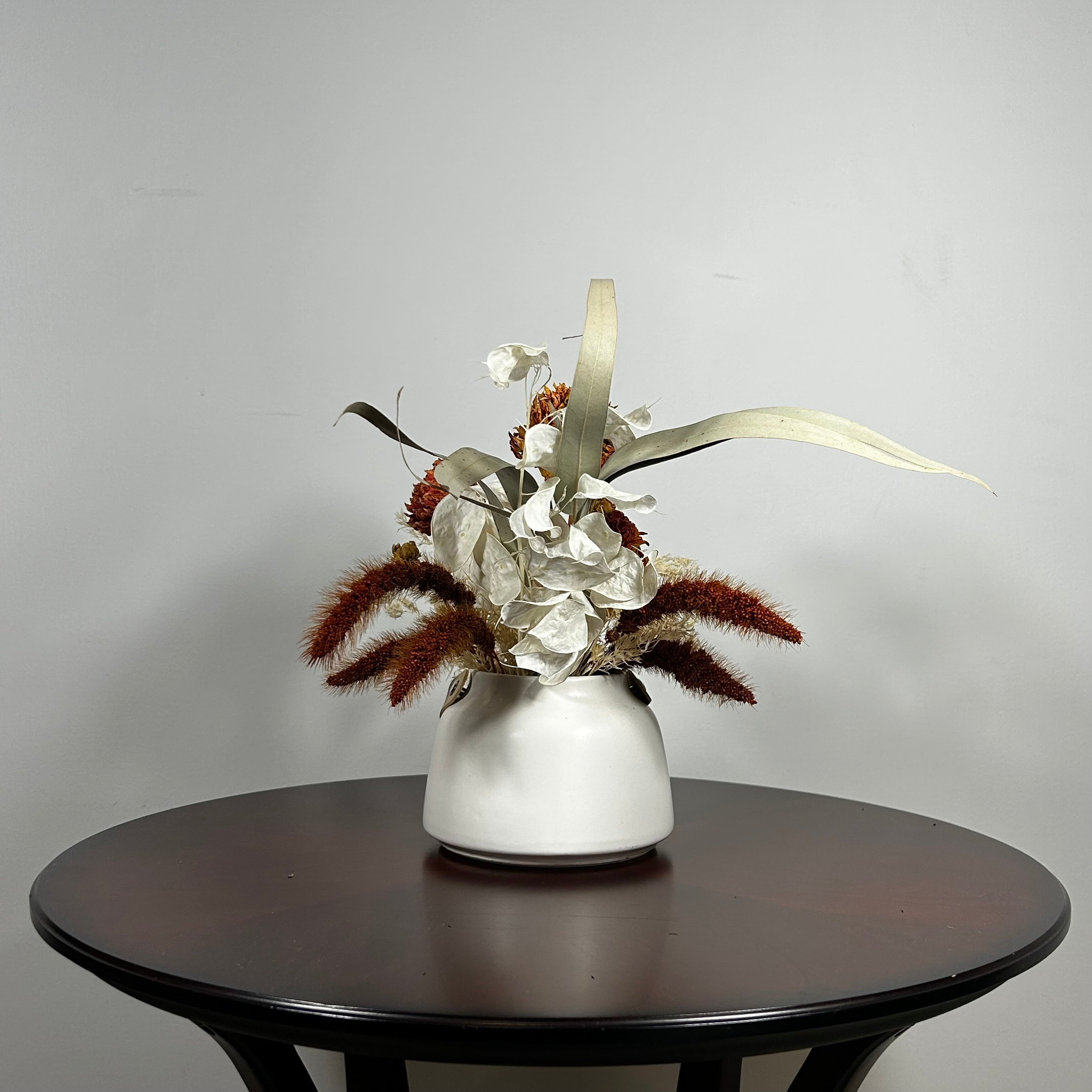 Dried flower arrangement with slender stems of natural green willow eucalyptus reach out from one side, complemented on the opposite side by the soft, bleached plumes of Calamgrostis canadensis. Ethereal Lunaria in pristine white adds a delicate, luminous quality. The featured floral is a cluster of five Dahlia stems, their petals a beautiful rust tone that gracefully fades into a vibrant golden heart, a perfect echo of the setting sun.